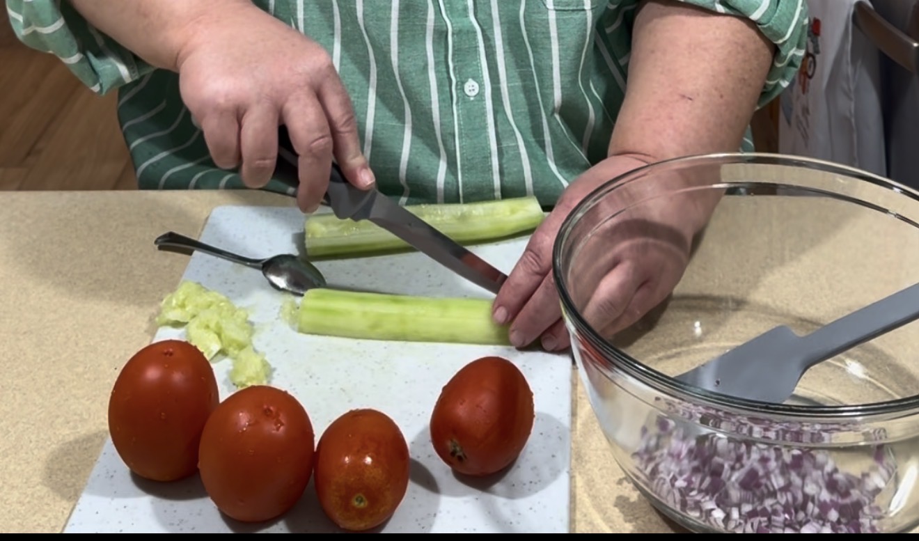 A person in a green striped shirt slices a peeled cucumber on a cutting board with a knife. Nearby are chopped celery, four tomatoes, and a bowl of diced red onion with a gray spatula.