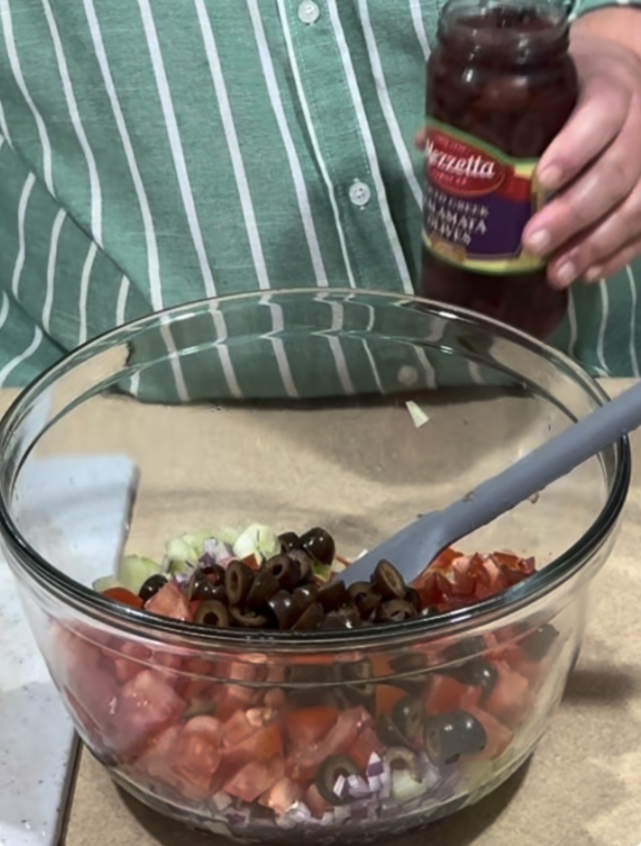 A person in a green striped shirt holds a jar of Mezzetta black olives above a glass bowl filled with chopped tomatoes, onions, celery, and olives. A spoon rests in the bowl.