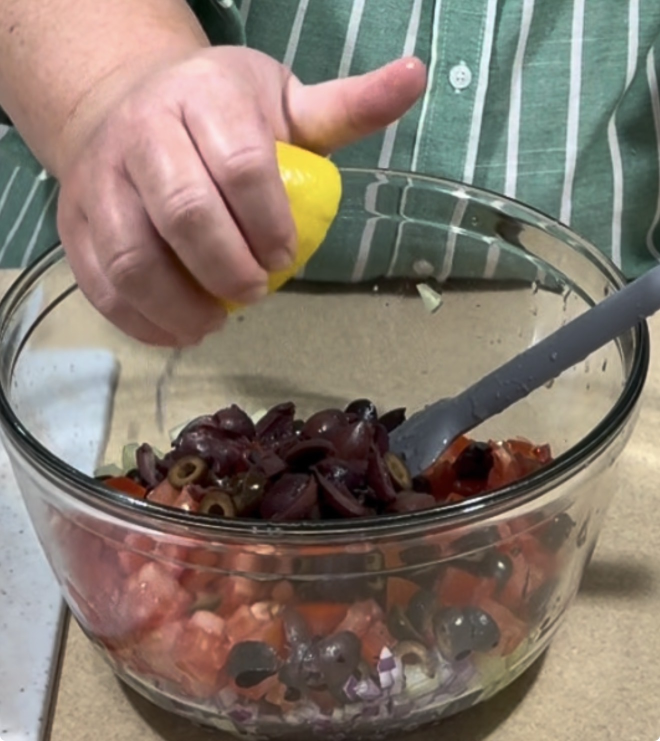 A person squeezes a lemon over a glass bowl filled with chopped tomatoes, olives, and onions. A gray spoon rests in the bowl. The person wears a green and white striped shirt.