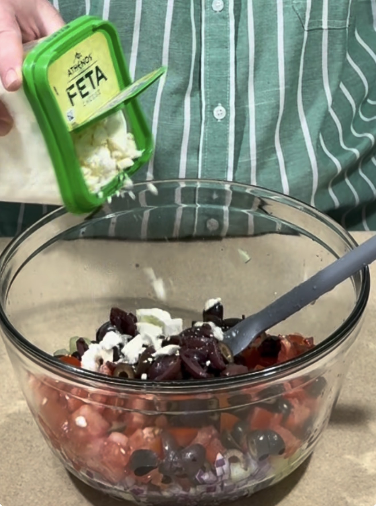 A person in a green striped shirt sprinkles crumbled feta cheese from a green container into a glass bowl filled with chopped vegetables and olives.