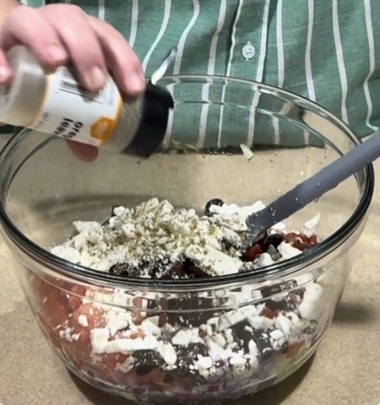 A person in a green striped shirt sprinkles seasoning from a bottle into a glass bowl filled with chopped vegetables and crumbled cheese, preparing a salad.