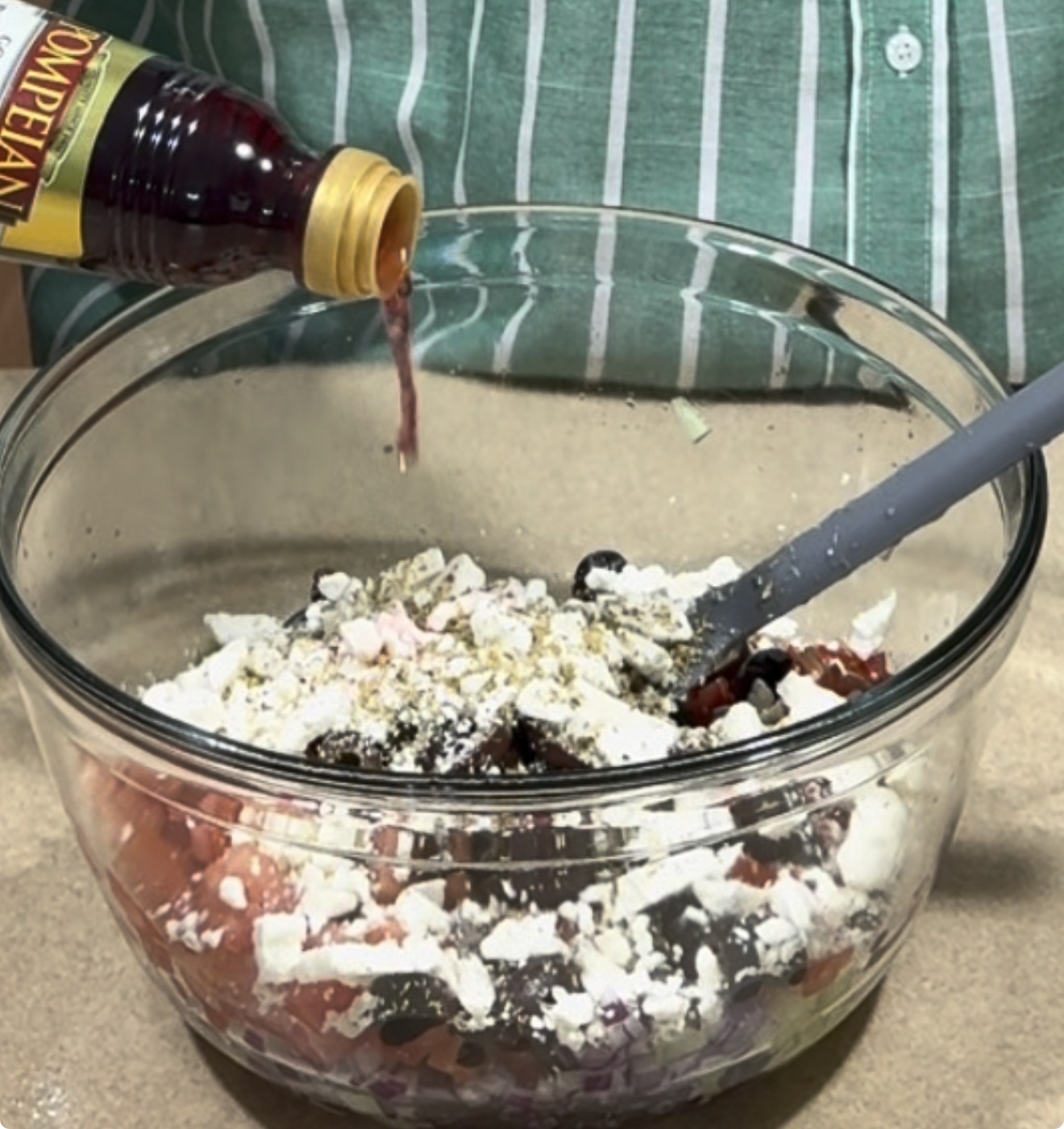 A person pours red wine vinegar from a bottle into a glass bowl filled with chopped vegetables, olives, and crumbled feta cheese, mixing ingredients for a salad.