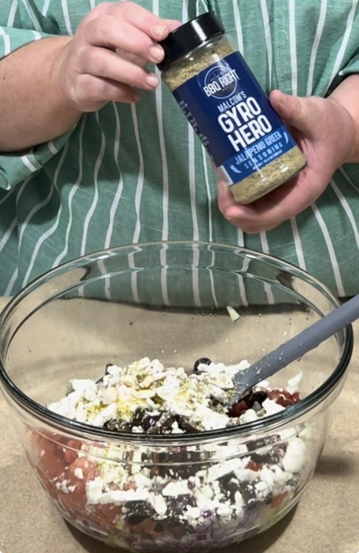 A person in a green striped shirt holds a bottle of Gyro Hero seasoning above a glass bowl filled with ingredients like feta cheese, olives, onions, and tomatoes, preparing a dish.