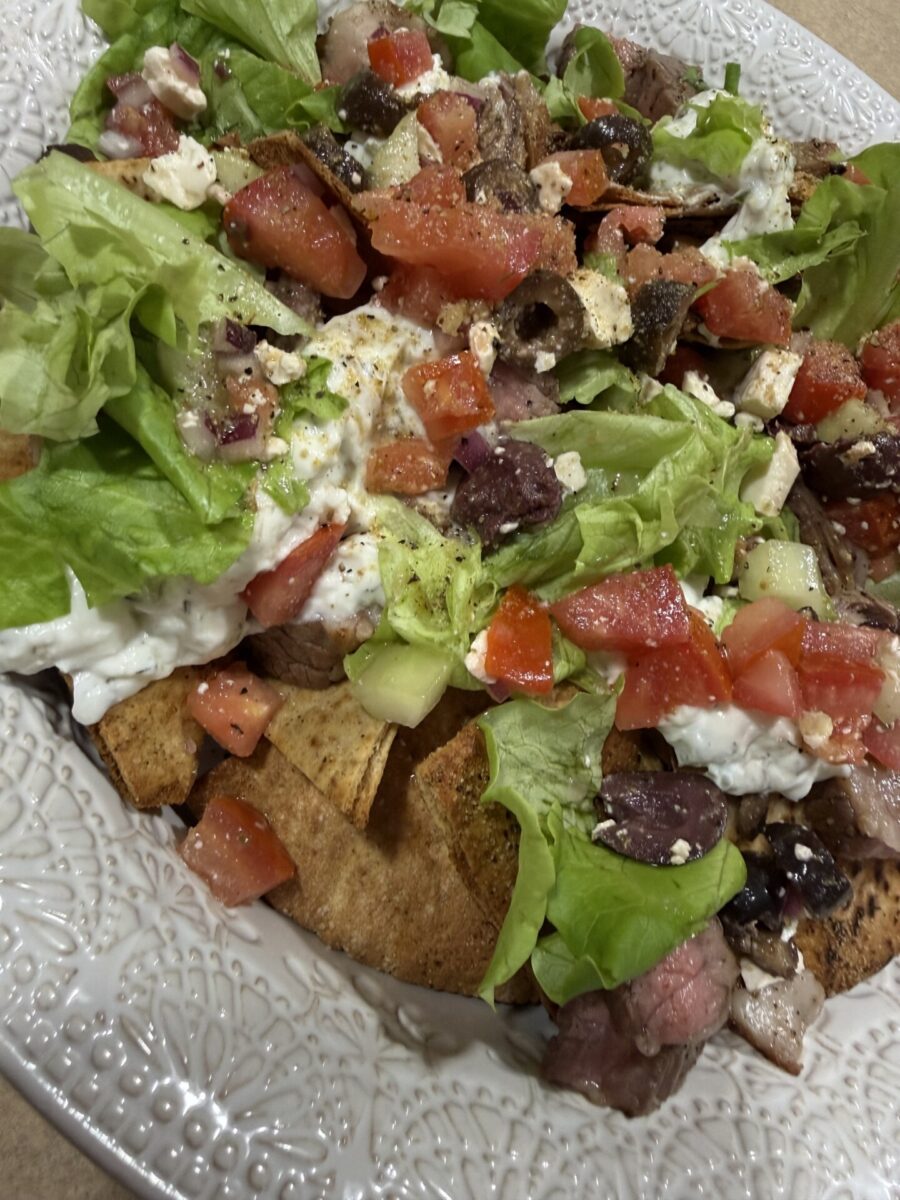 A close-up of a plate with pita chips topped with lettuce, diced tomatoes, cucumbers, olives, feta cheese, and tzatziki sauce, served on a white decorative dish.