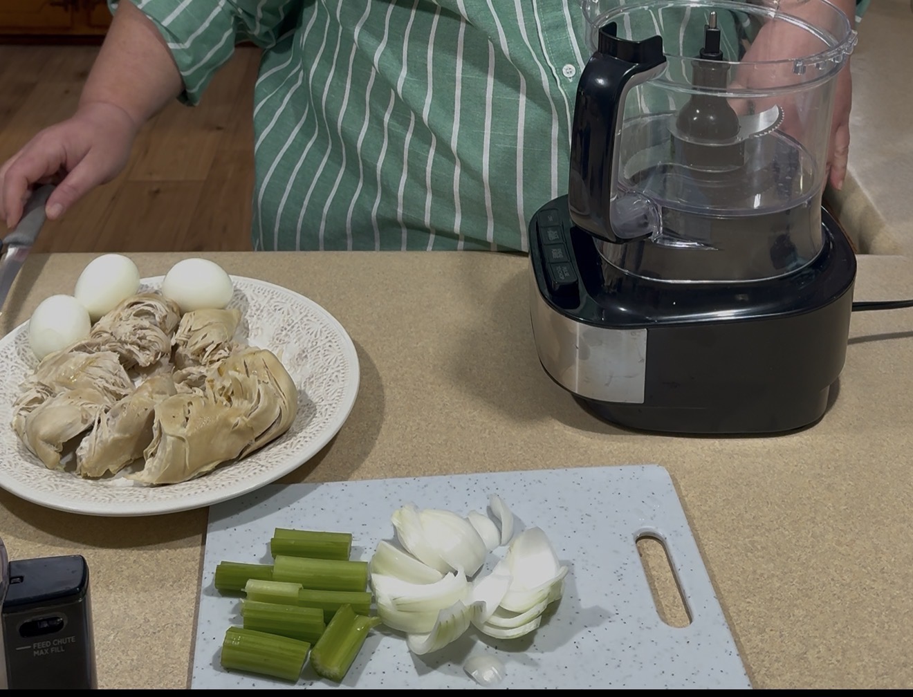 A person in a green striped shirt stands by a kitchen counter with a food processor. Nearby are a plate with cooked chicken and boiled eggs, and a cutting board with chopped celery and onions.