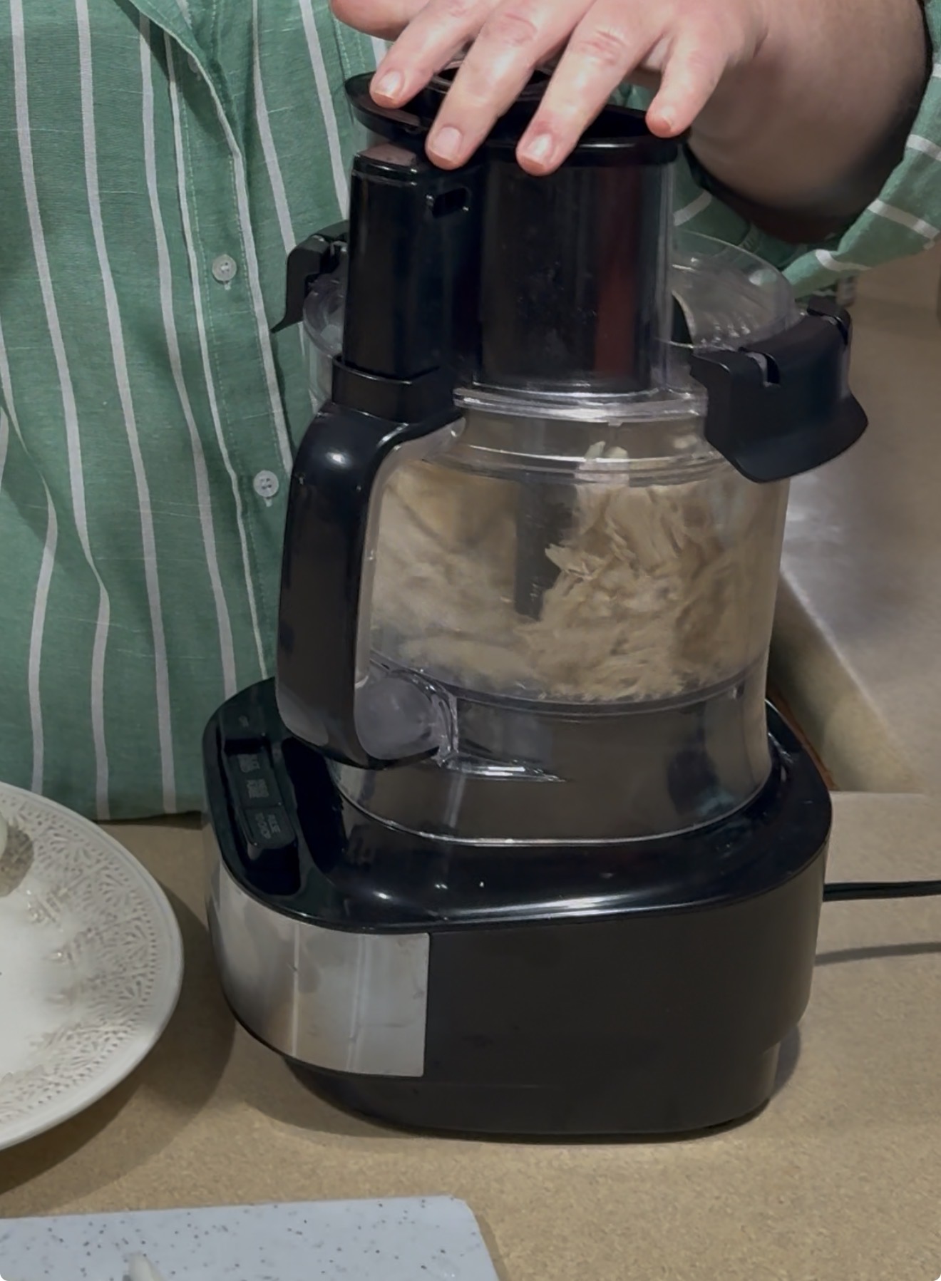 A person in a green and white striped shirt uses a food processor filled with shredded chicken, with a decorative plate nearby on the counter.