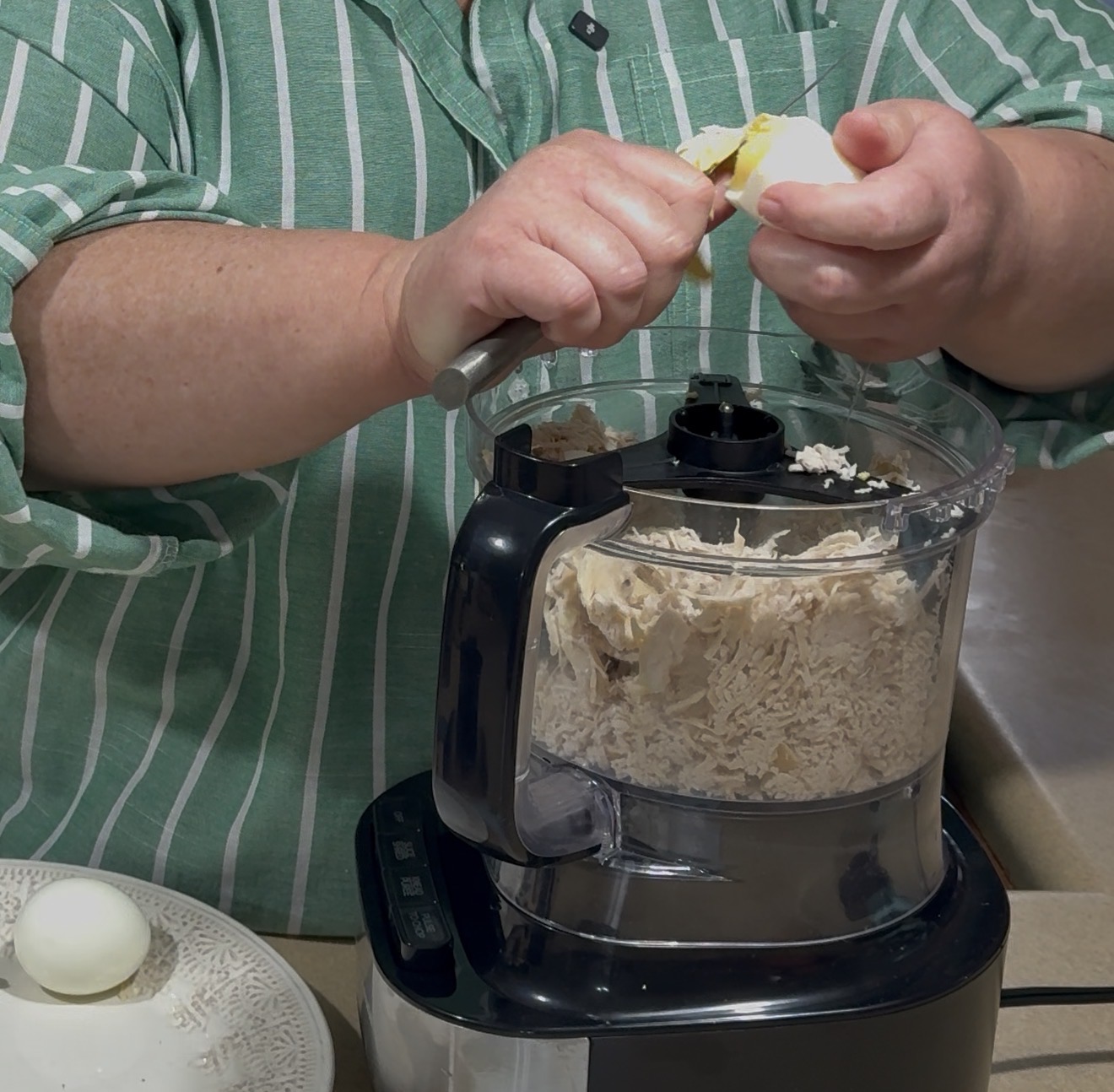 A person in a green striped shirt peels a hard-boiled egg over a food processor filled with shredded chicken. A plate with eggs is on the counter nearby.