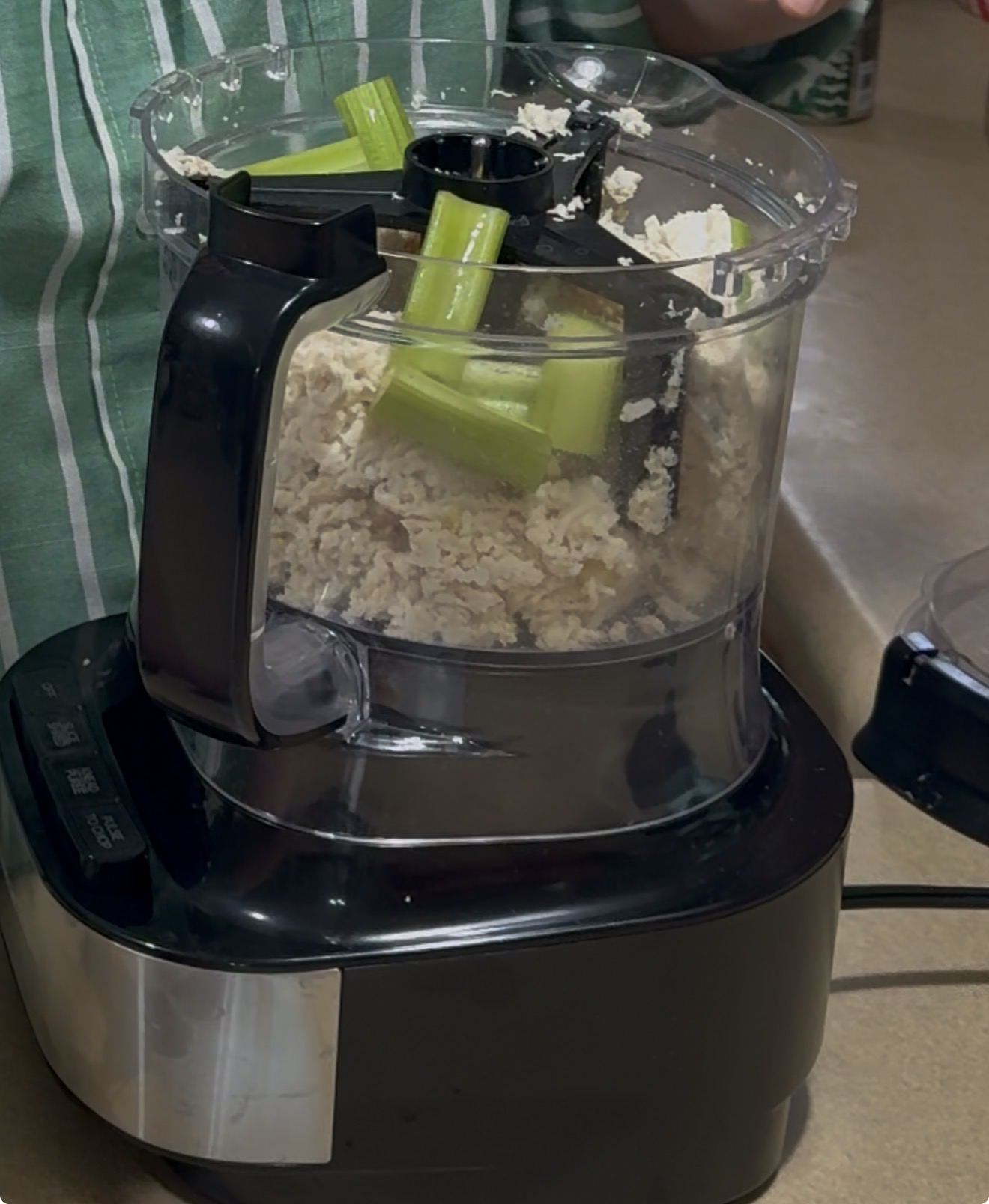 A food processor on a countertop with chunks of celery and chopped tofu inside. A person in a green and white striped shirt stands behind the appliance, with their hand visible at the top right.