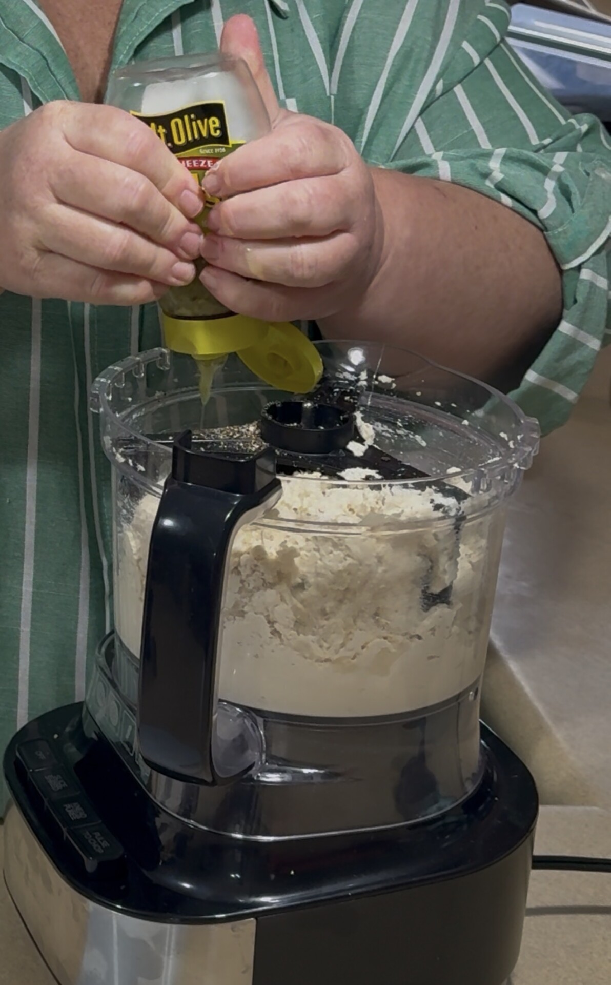 A person in a green striped shirt squeezes a bottle of olive oil into a food processor filled with flour mixture on a kitchen countertop.