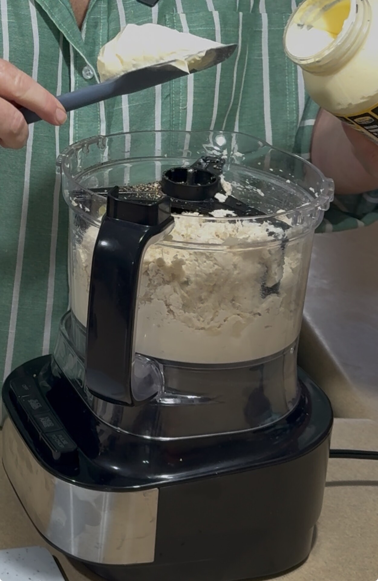 A person in a green striped shirt adds mayonnaise from a jar into a food processor filled with dough or a creamy mixture, using a spatula.