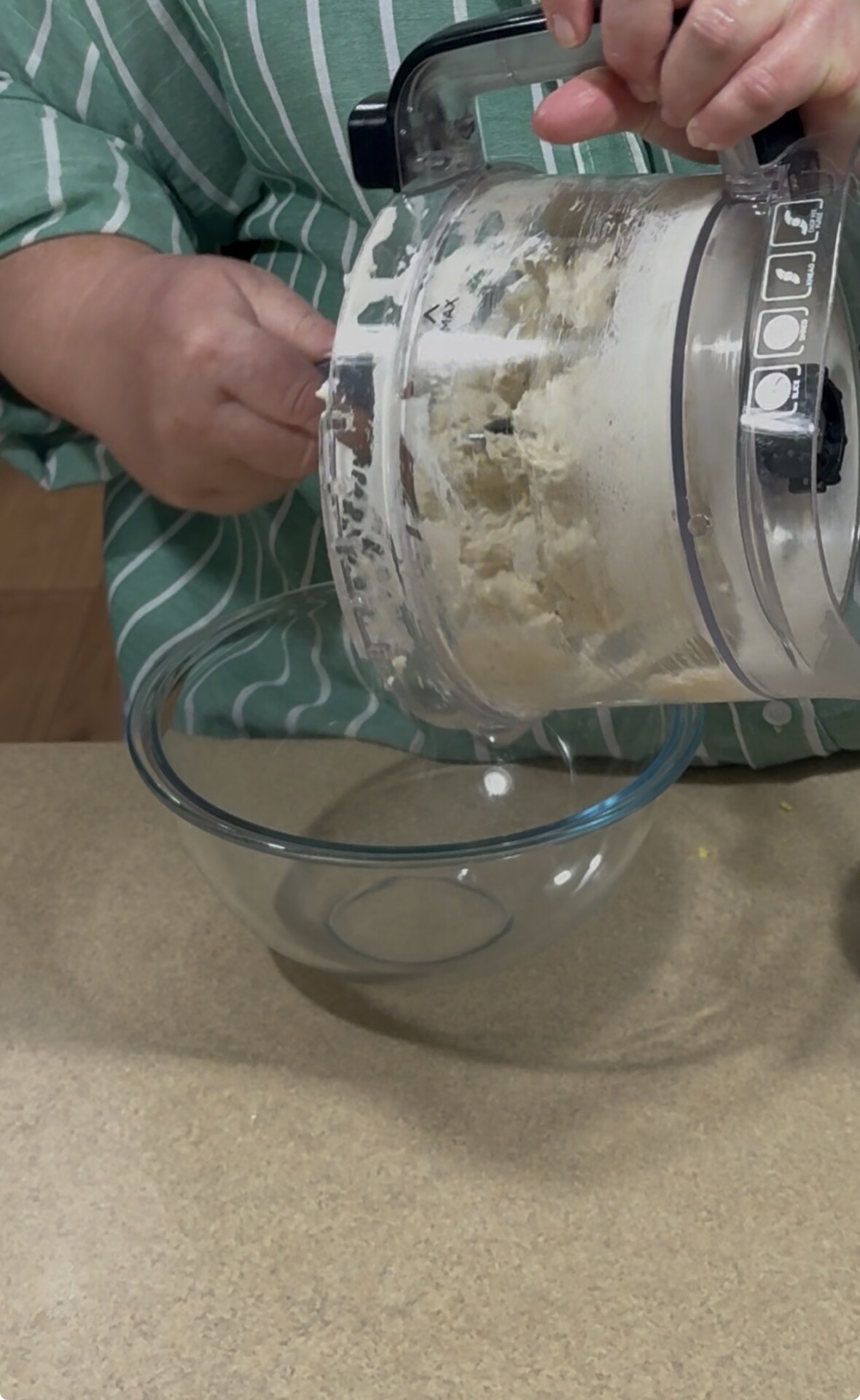A person in a green striped shirt is scraping dough from a food processor into a clear glass bowl on a kitchen counter.
