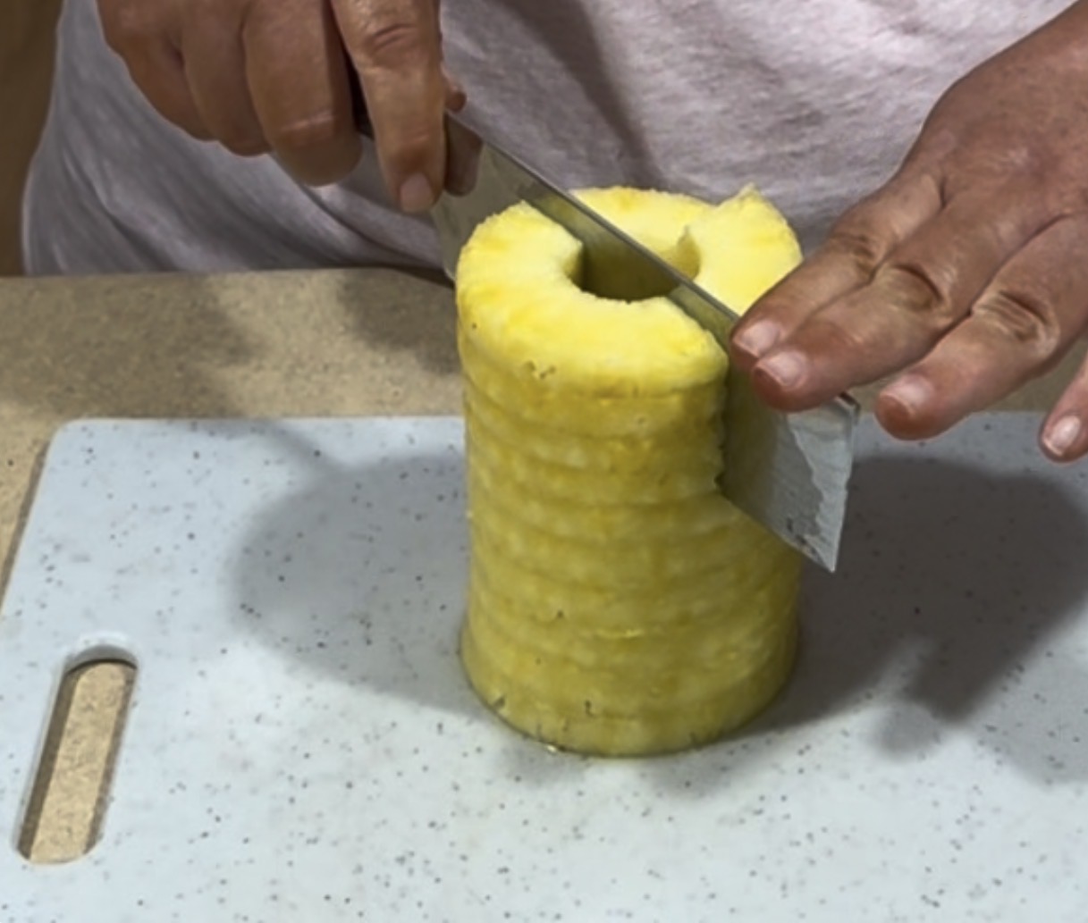 A person slices a cored, peeled pineapple on a white cutting board. The pineapple is standing upright and the person is holding it steady while cutting with a large knife.