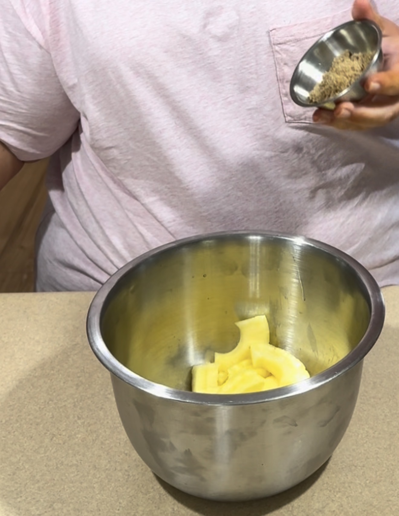 A person in a pink shirt holds a small bowl of brown sugar above a metal mixing bowl containing chunks of butter, positioned on a beige countertop.