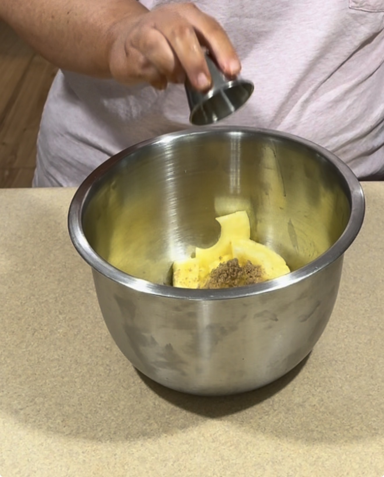 A person adds spices from a small metal cup into a large metal bowl containing chopped pineapple pieces on a kitchen counter.
