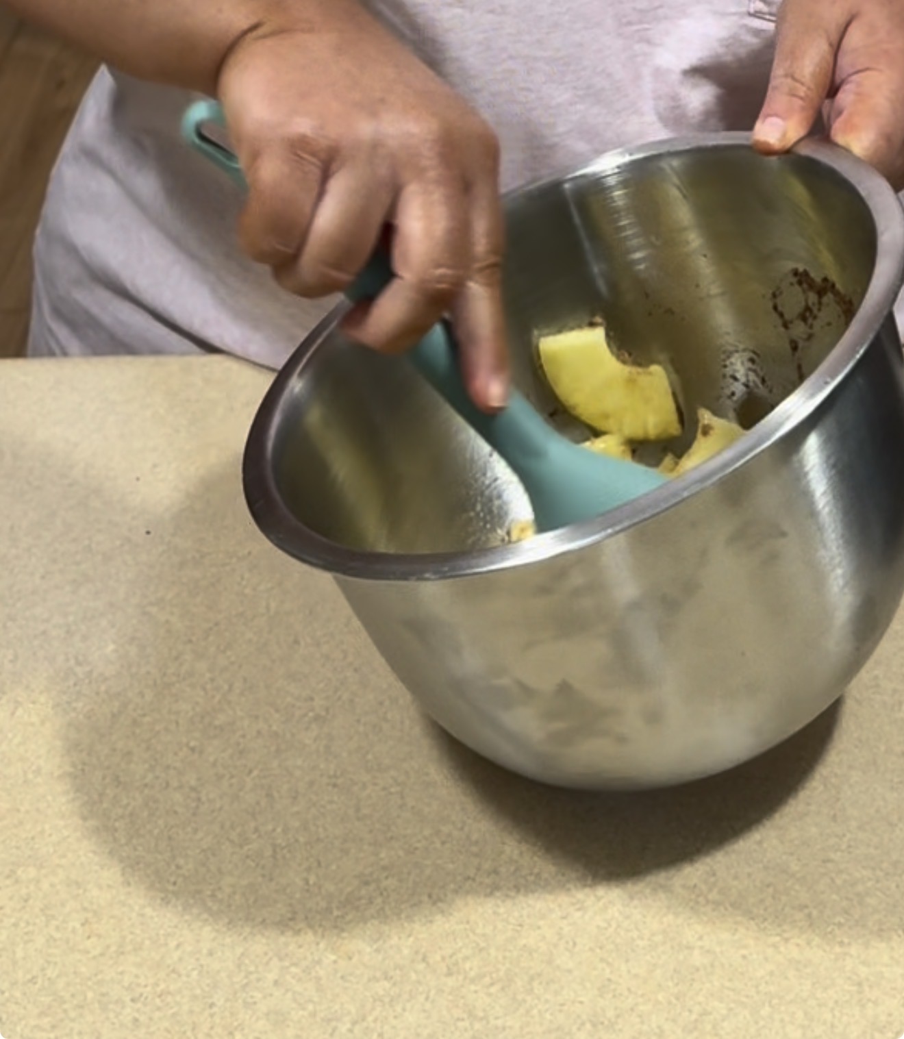 A person uses a blue spatula to mix pineapple pieces in a large stainless steel bowl on a kitchen counter.