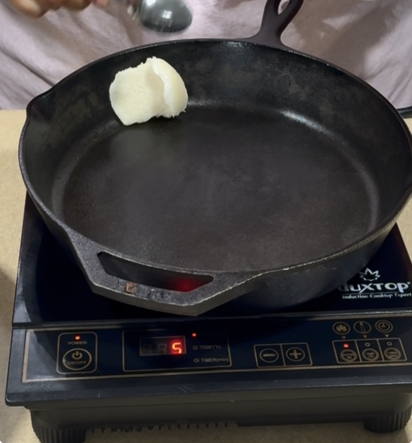 A person adds a spoonful of solid white fat, likely shortening or lard, to a black cast iron skillet heating on an electric cooktop set to level 5.