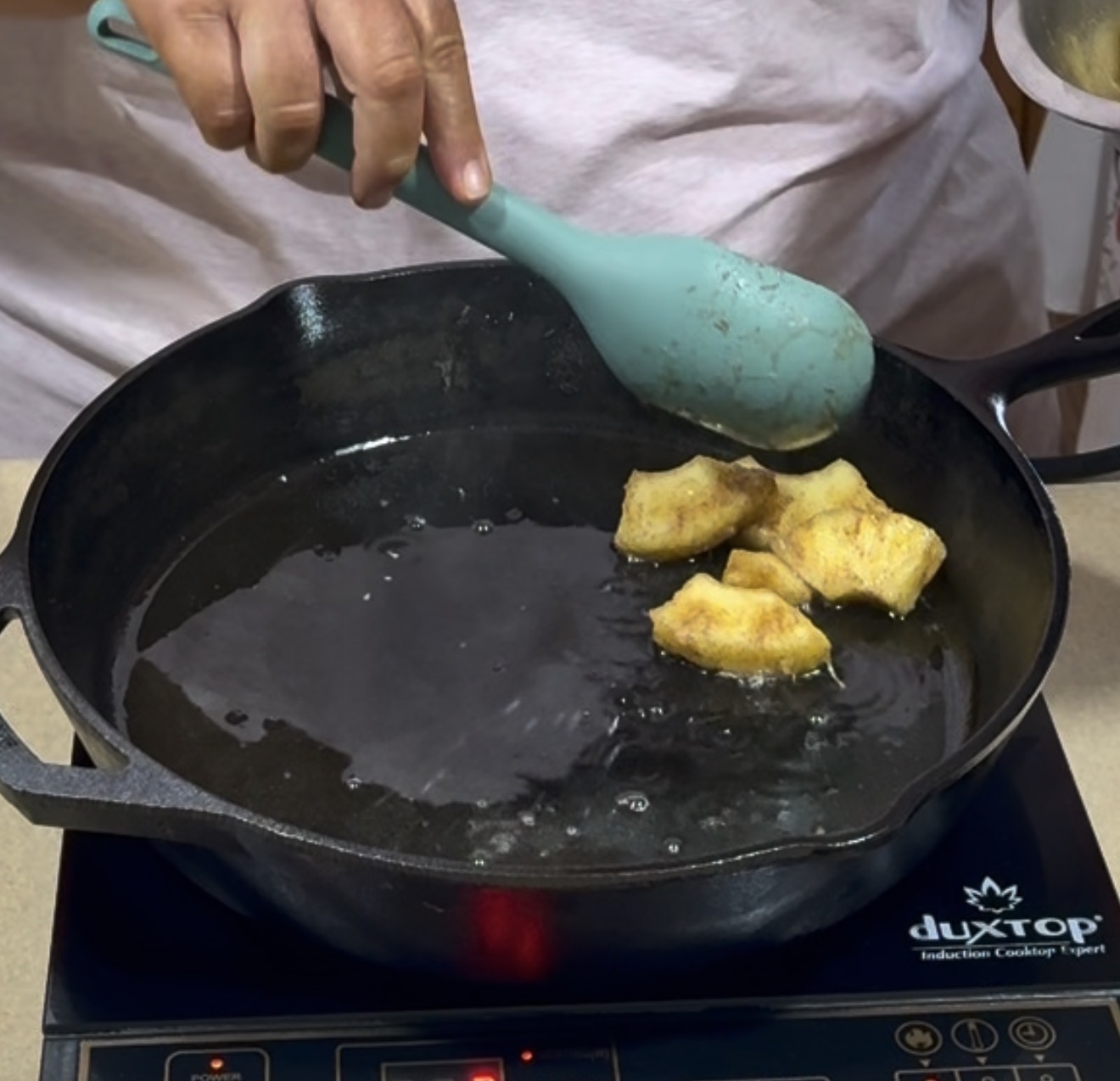 A person uses a blue spatula to fry pieces of plantain in a black cast iron skillet on an induction cooktop. The skillet contains hot oil and the cooktop brand duxtop is visible.