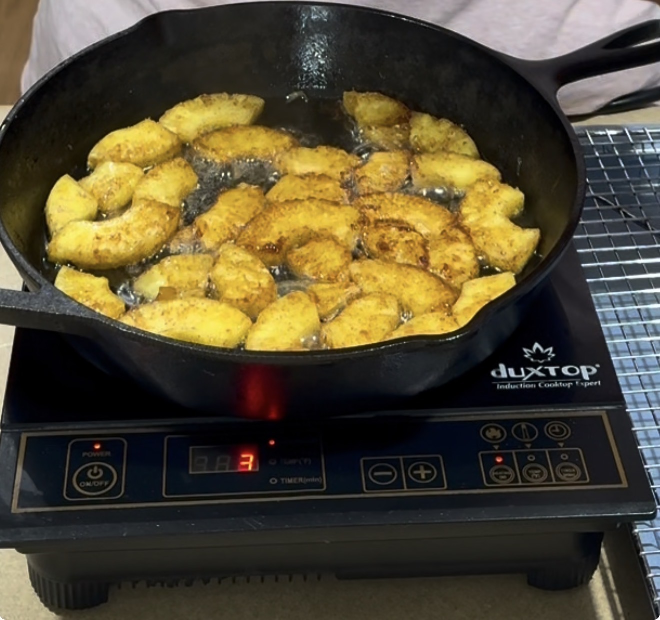 A cast iron skillet filled with frying, golden-brown battered pieces sits on an induction cooktop set to level 3, with a cooling rack nearby.