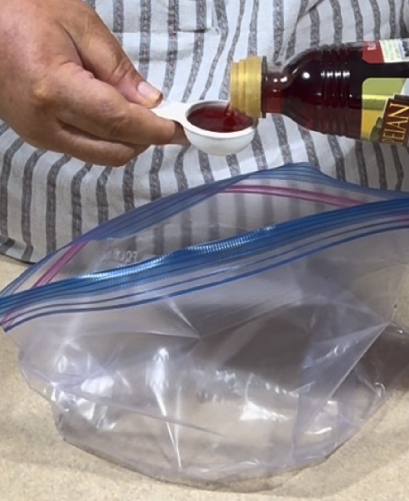 A person pours a reddish liquid from a bottle into a measuring spoon over an open, empty zip-top plastic bag placed on a countertop.