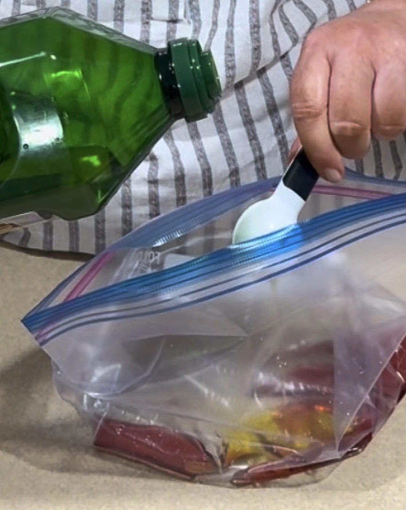 A person pours olive oil from a green bottle into a plastic zipper bag filled with food, using a white measuring spoon.