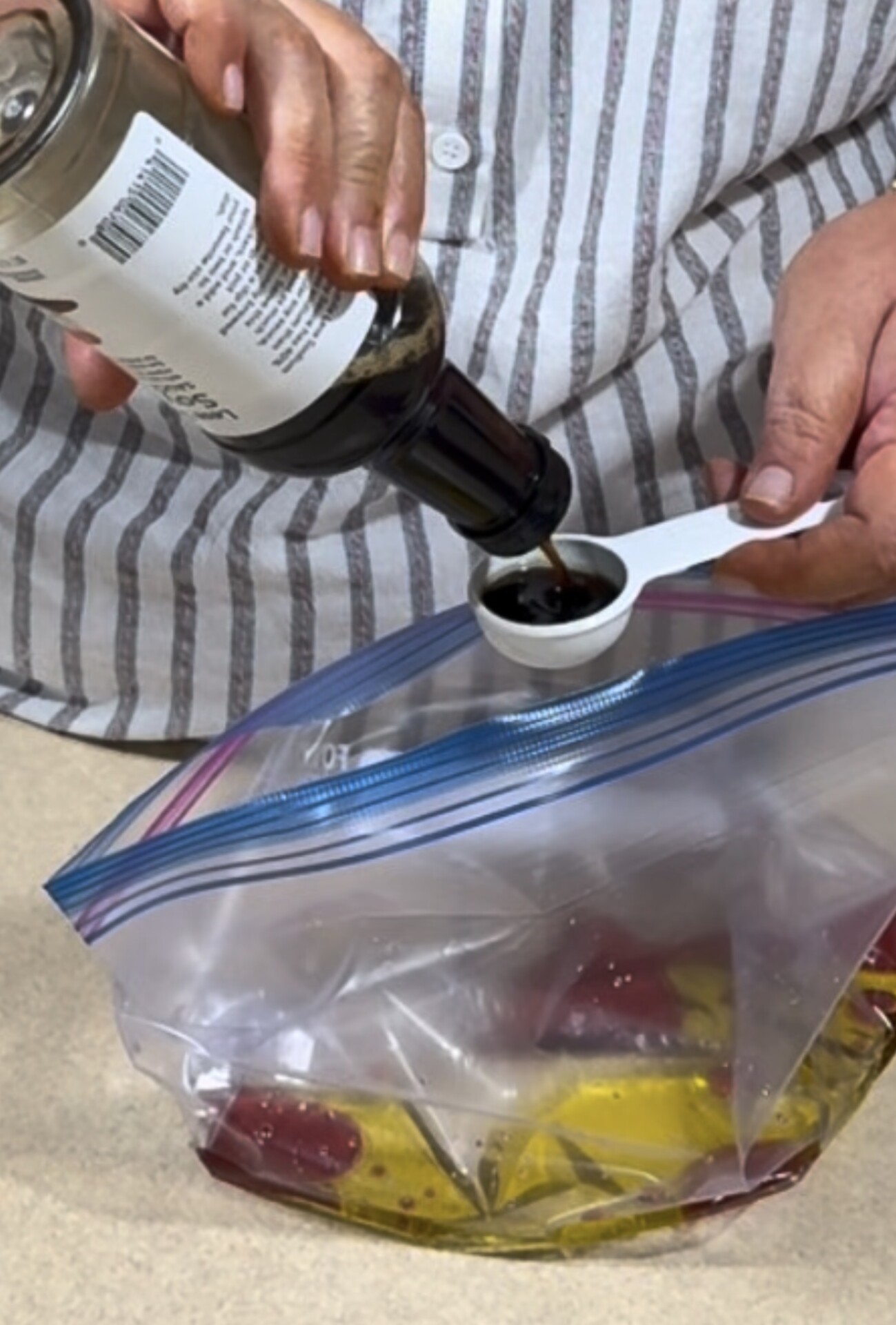 A person pours a dark liquid from a bottle into a white measuring spoon over a ziplock bag containing marinating food. The person is wearing a striped shirt.
