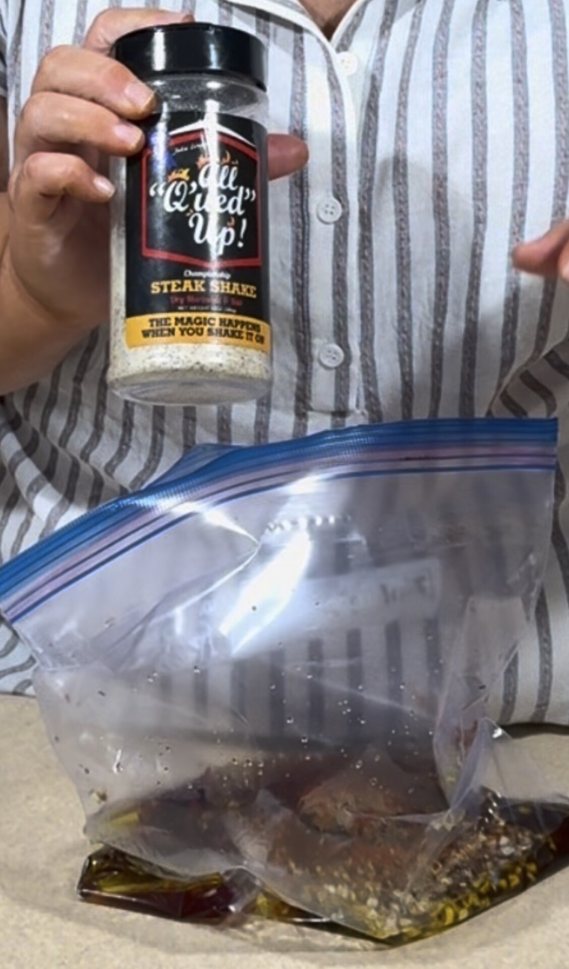 A person holds a container of steak seasoning above a ziplock bag filled with marinade and meat on a counter.