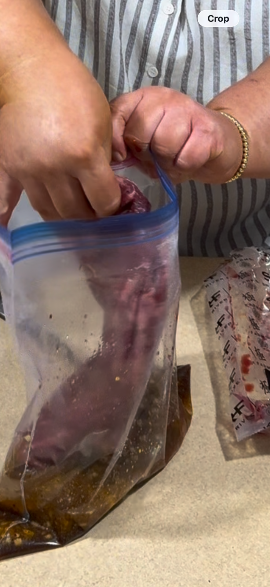 A person wearing a striped shirt places a piece of raw meat into a ziplock bag filled with marinade on a countertop. An unopened package of meat sits beside the bag.