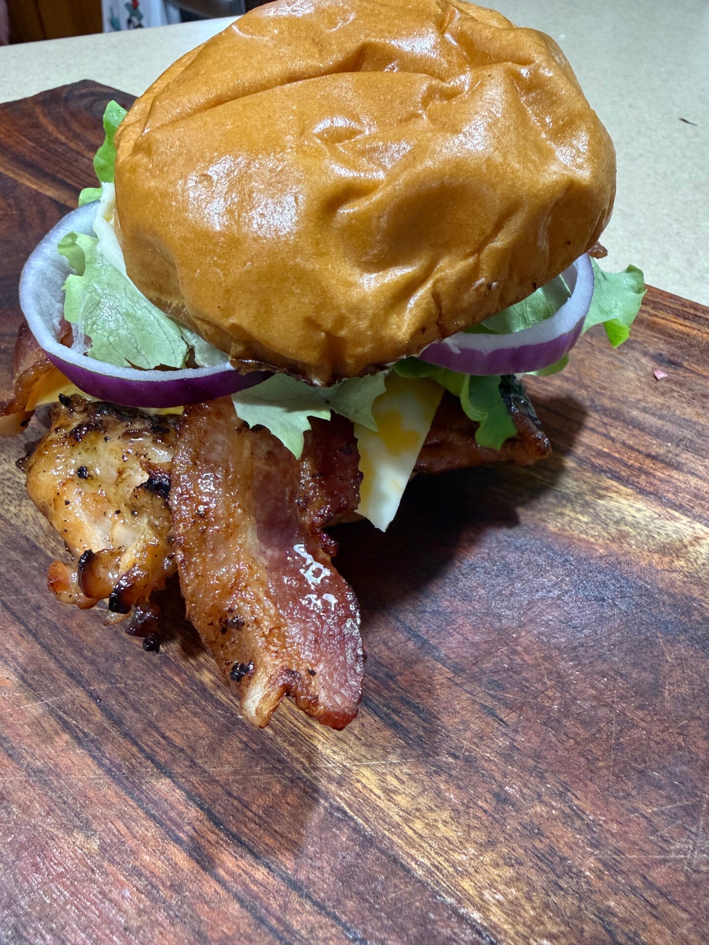 A close-up of a sandwich on a wooden cutting board, featuring crispy bacon, grilled chicken, cheese, lettuce, red onion, and a shiny brioche bun.