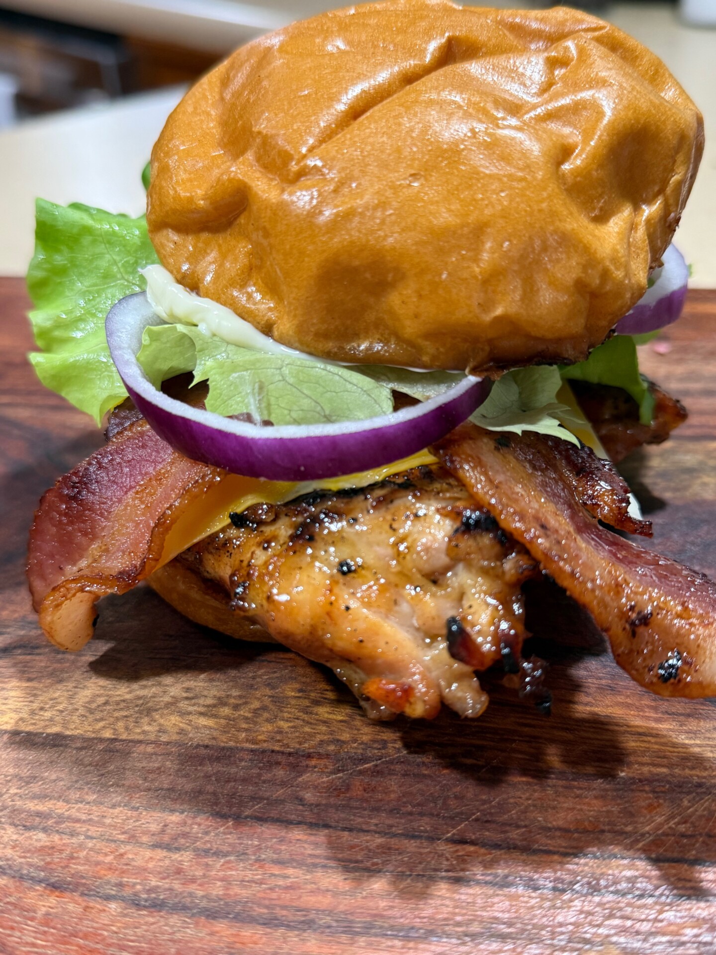 A close-up of a sandwich on a wooden surface, featuring grilled chicken, crispy bacon, red onion slices, lettuce, cheddar cheese, and a soft toasted bun.
