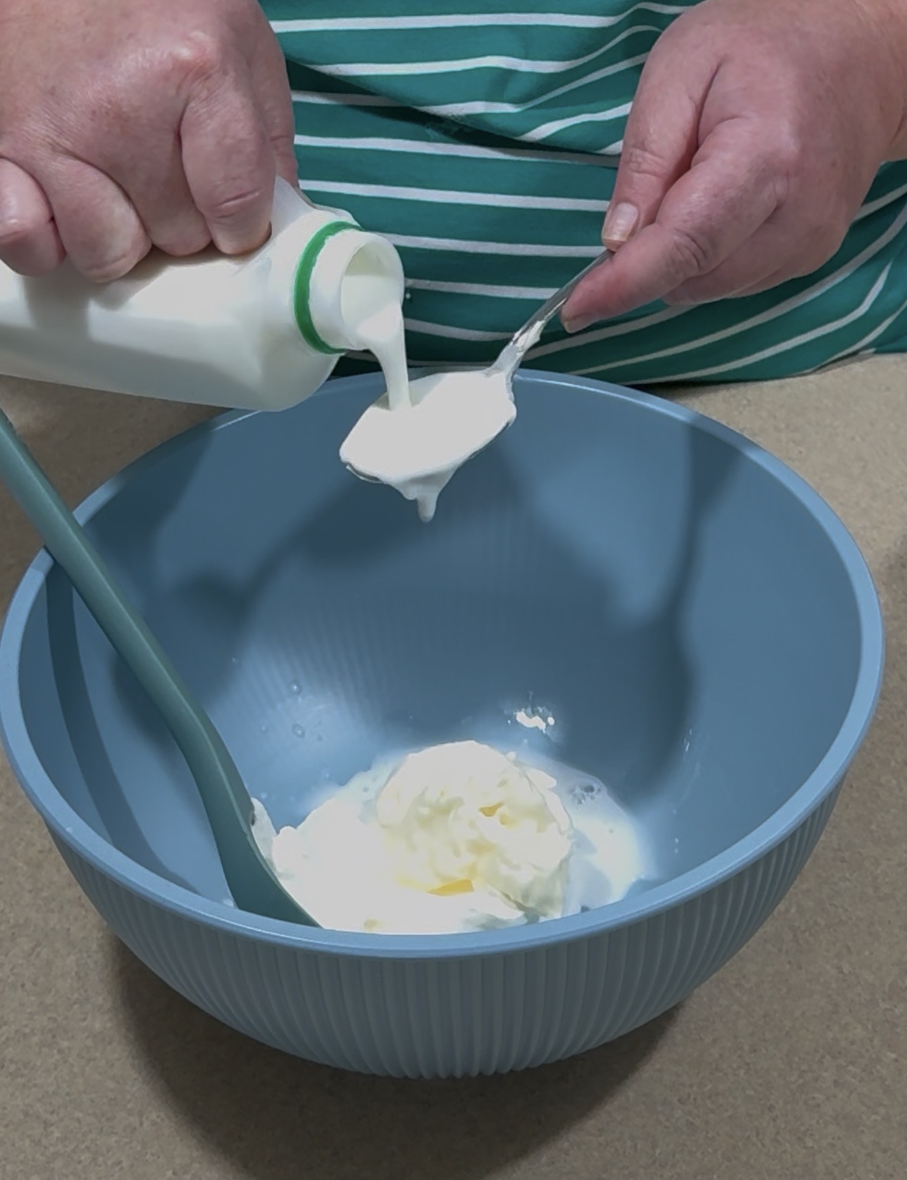 A person pours milk from a bottle into a spoon over a blue mixing bowl containing cream or yogurt, next to a blue spatula, on a kitchen countertop.