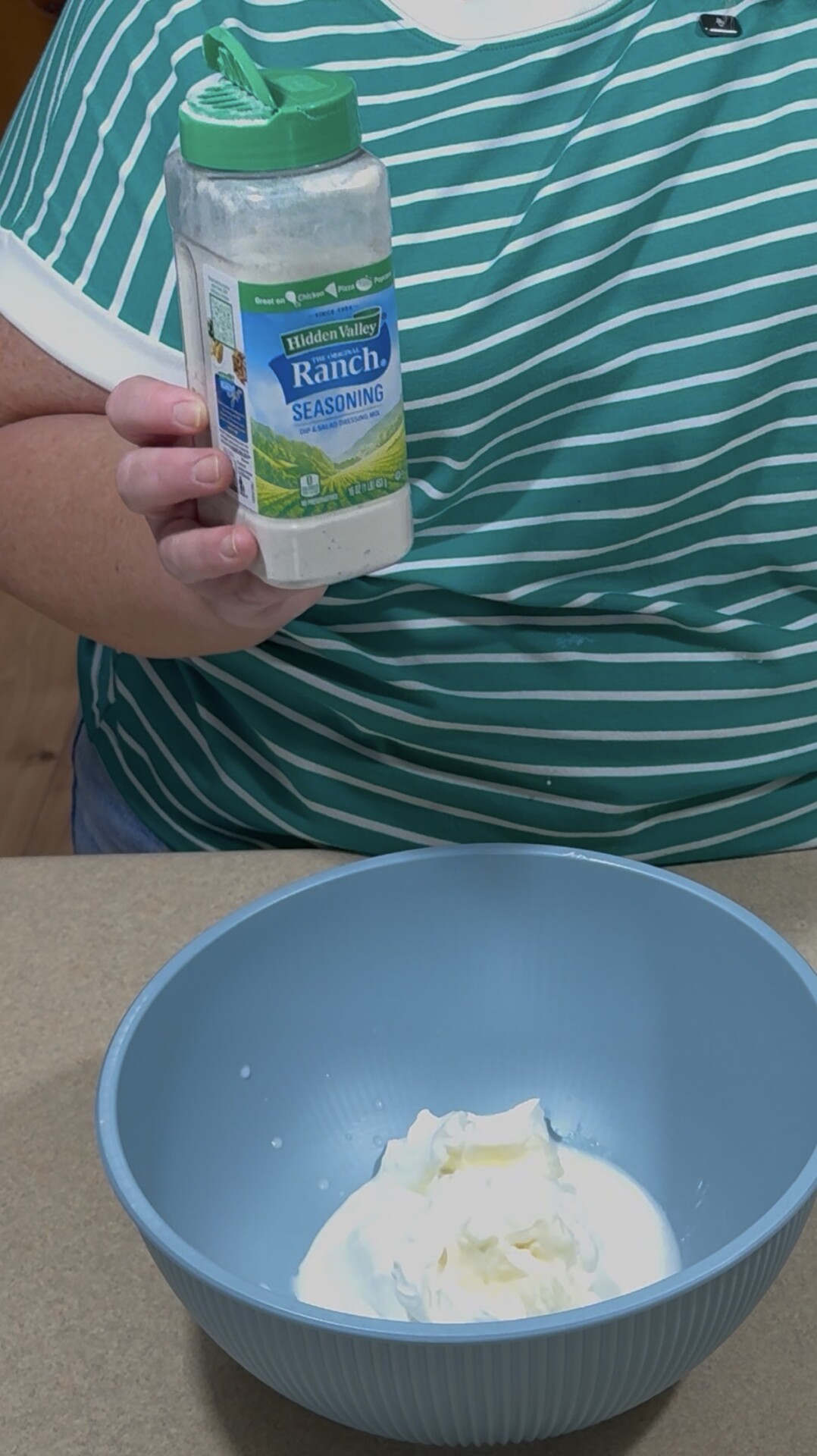 A person in a green striped shirt holds a container of Hidden Valley Ranch seasoning over a blue bowl with white ingredients, possibly mayonnaise and sour cream.