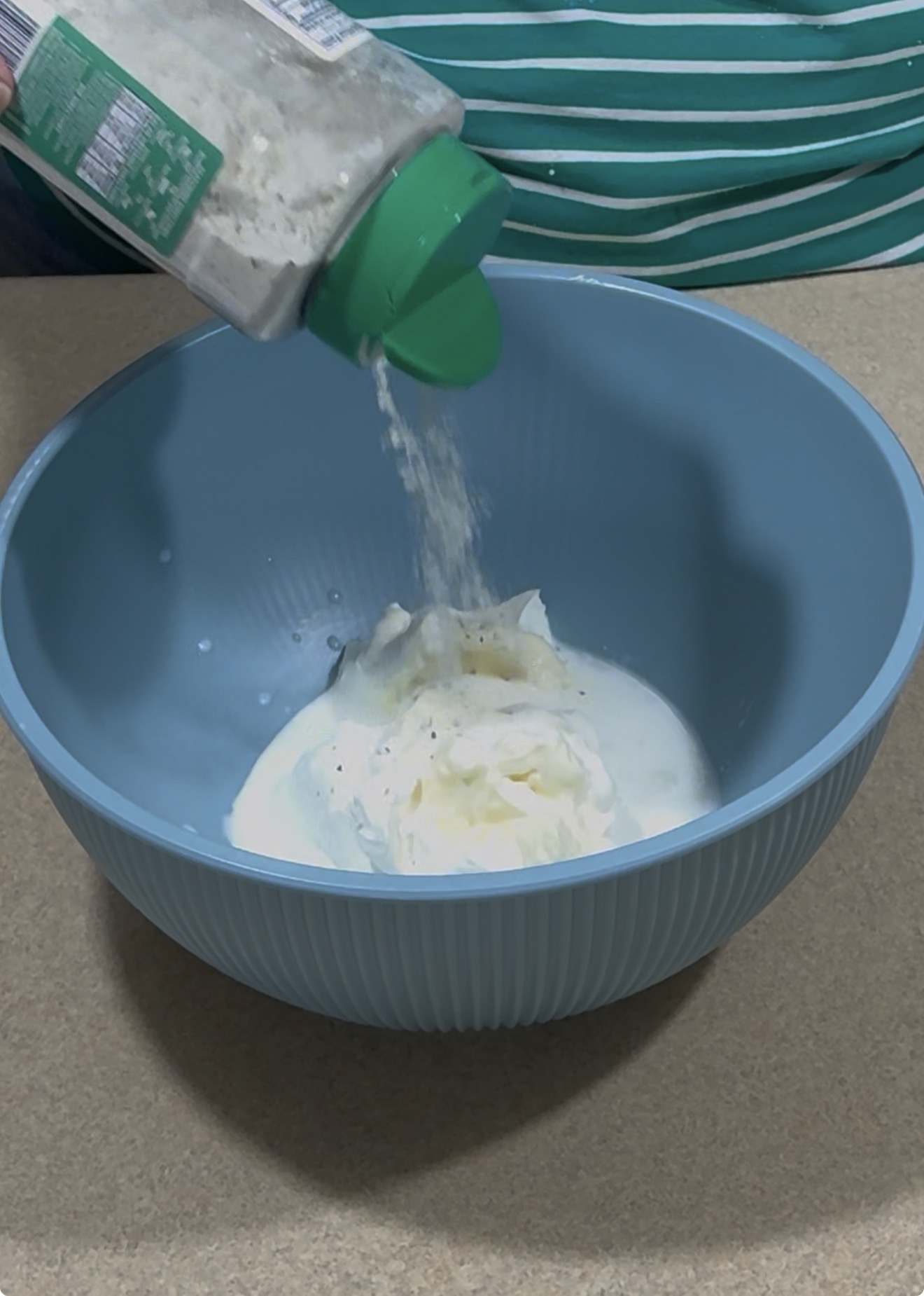 A person pours seasoning from a green shaker into a blue bowl containing mayonnaise and milk or cream, set on a beige countertop.