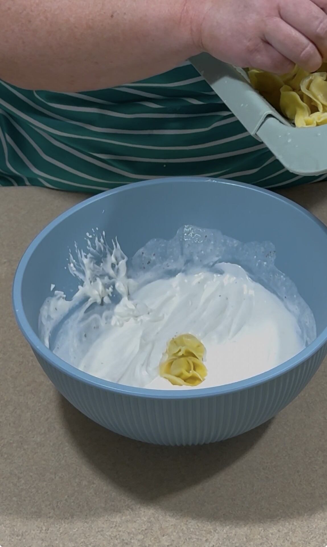 A person adds uncooked pasta to a blue bowl filled with thick white cream or sauce on a beige countertop.