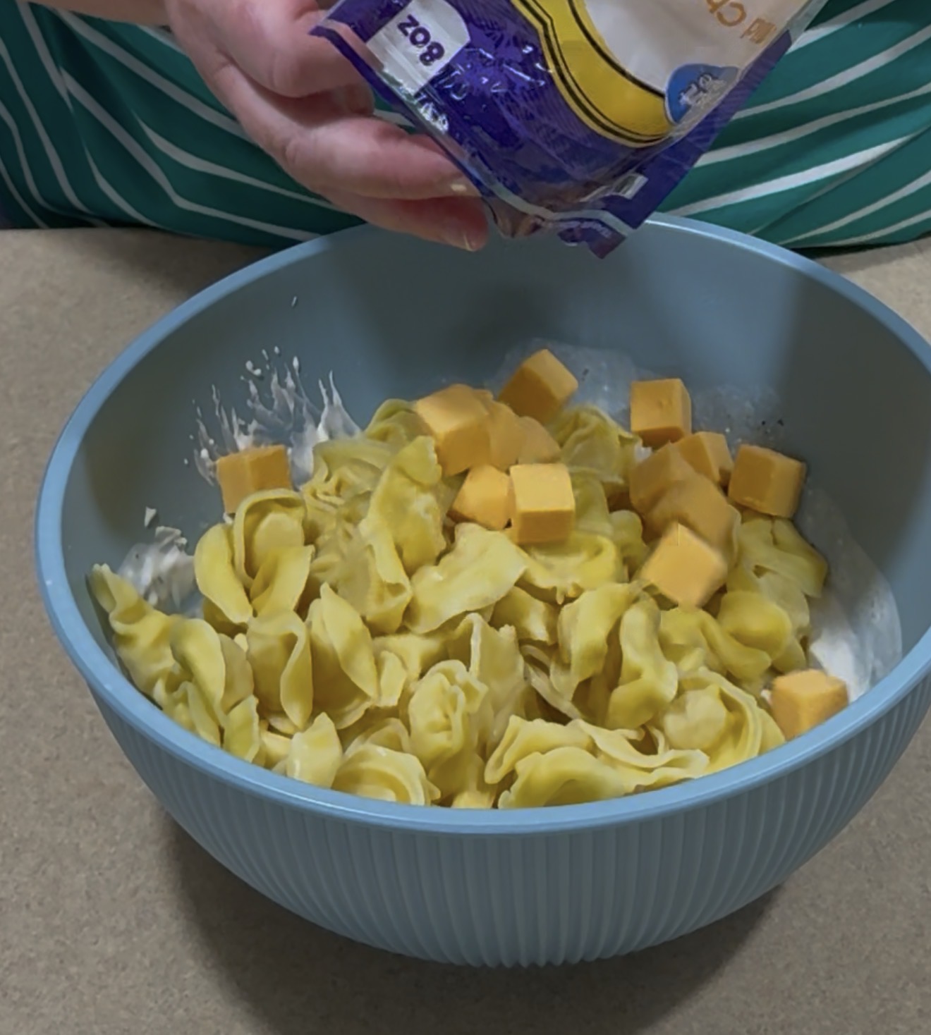 A person is preparing a dish by adding shredded cheese from a bag to a bowl filled with tortellini, cheese cubes, and a creamy white sauce.