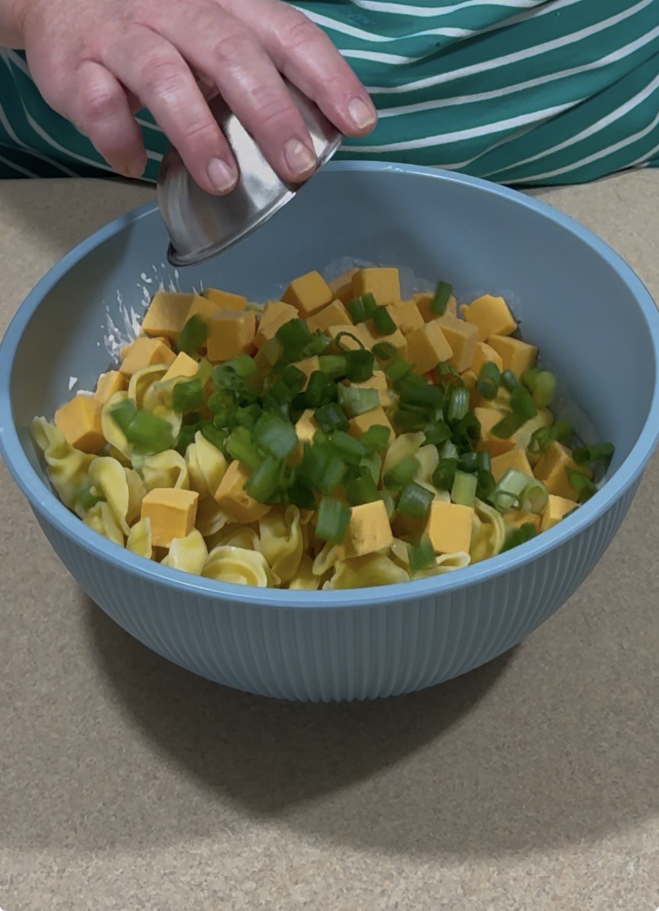 A person adds an ingredient from a small metal cup to a blue bowl containing tortellini, cubed cheddar cheese, and chopped green onions, on a beige countertop.