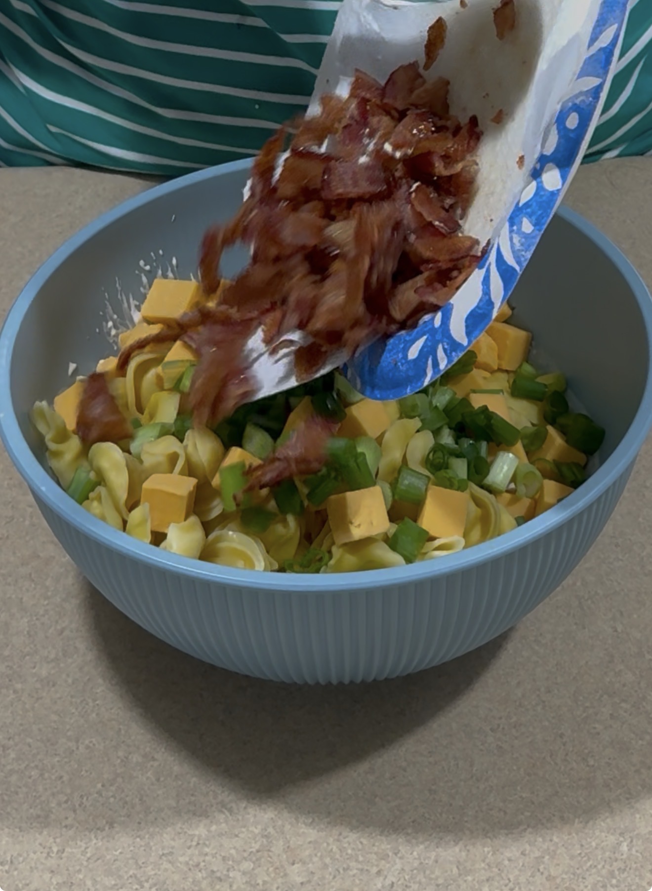 A blue bowl filled with uncooked pasta, cheese cubes, and chopped green onions sits on a counter as crispy bacon pieces are being added from a paper plate.
