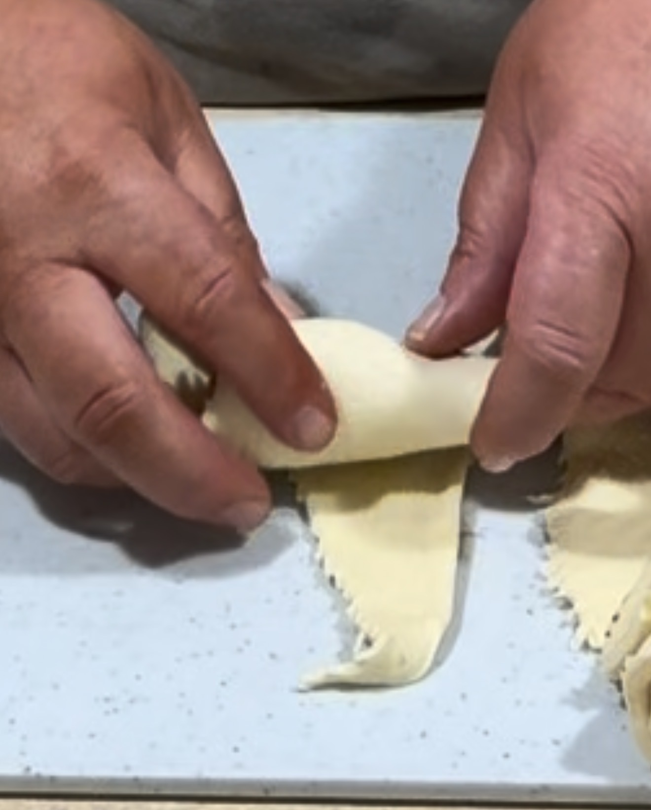Hands rolling a piece of dough into a crescent shape on a speckled white surface, with additional dough pieces visible nearby.