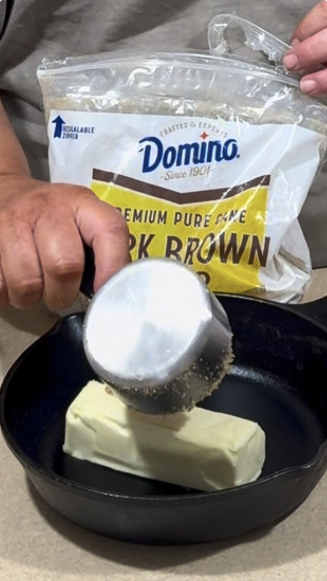 A person pours a cup of dark brown sugar from a Domino bag into a skillet with a stick of butter.