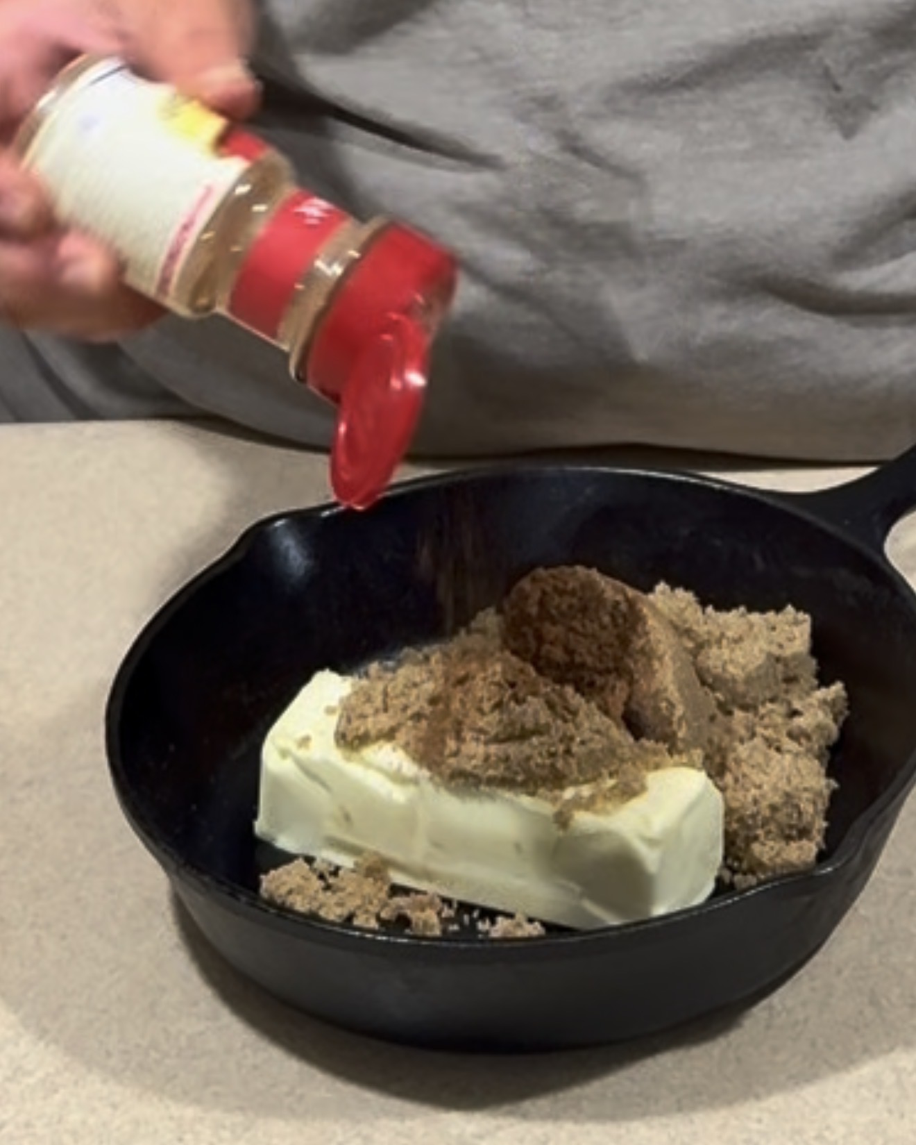 A person sprinkles seasoning from a bottle over a stick of butter and a pile of brown sugar in a black cast iron skillet on a kitchen counter.