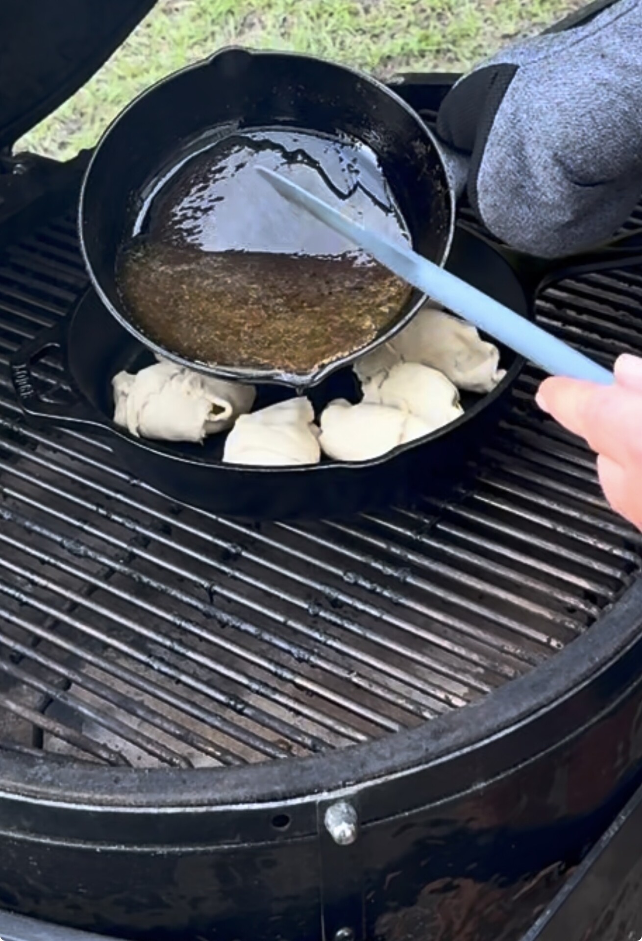 A person stirs liquid in a cast iron skillet on an outdoor grill. Several balls of dough are arranged in the same skillet. An oven mitt rests nearby. Grass is visible in the background.