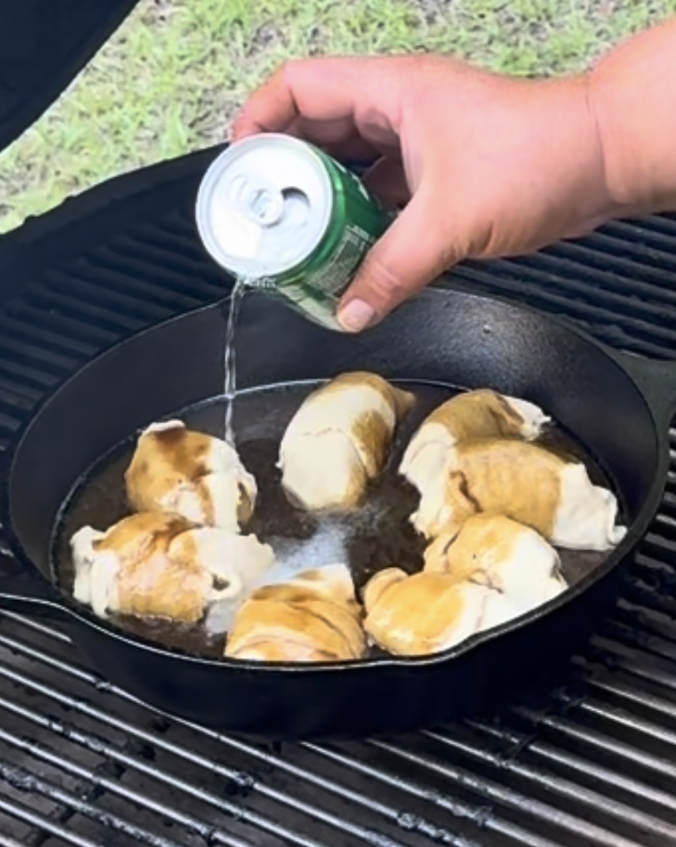 A hand pours a can of clear soda over crescent roll dough pieces in a cast iron skillet on an outdoor grill, with grass visible in the background.