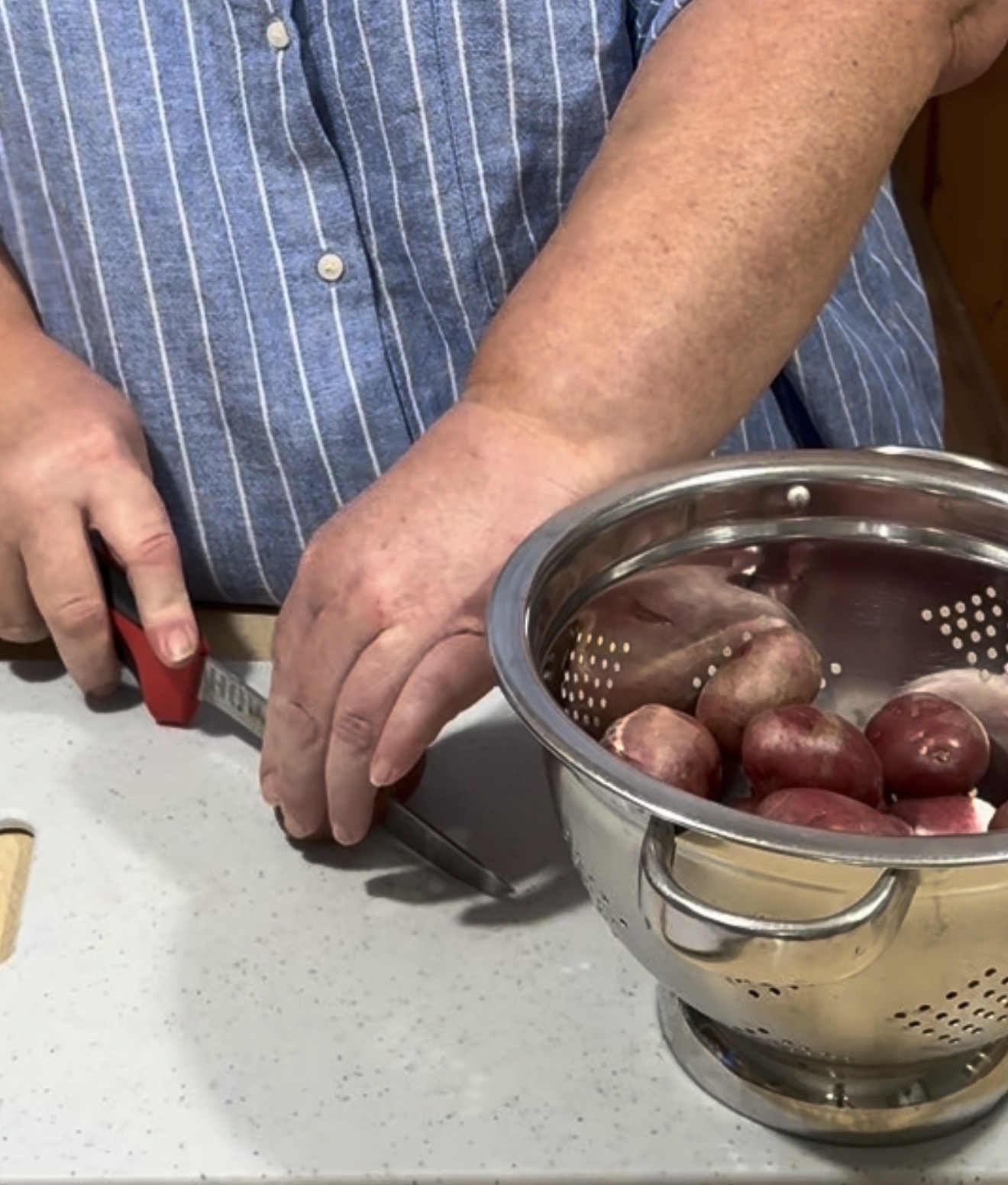 A person in a blue striped shirt slices potatoes on a countertop next to a metal colander filled with red potatoes.