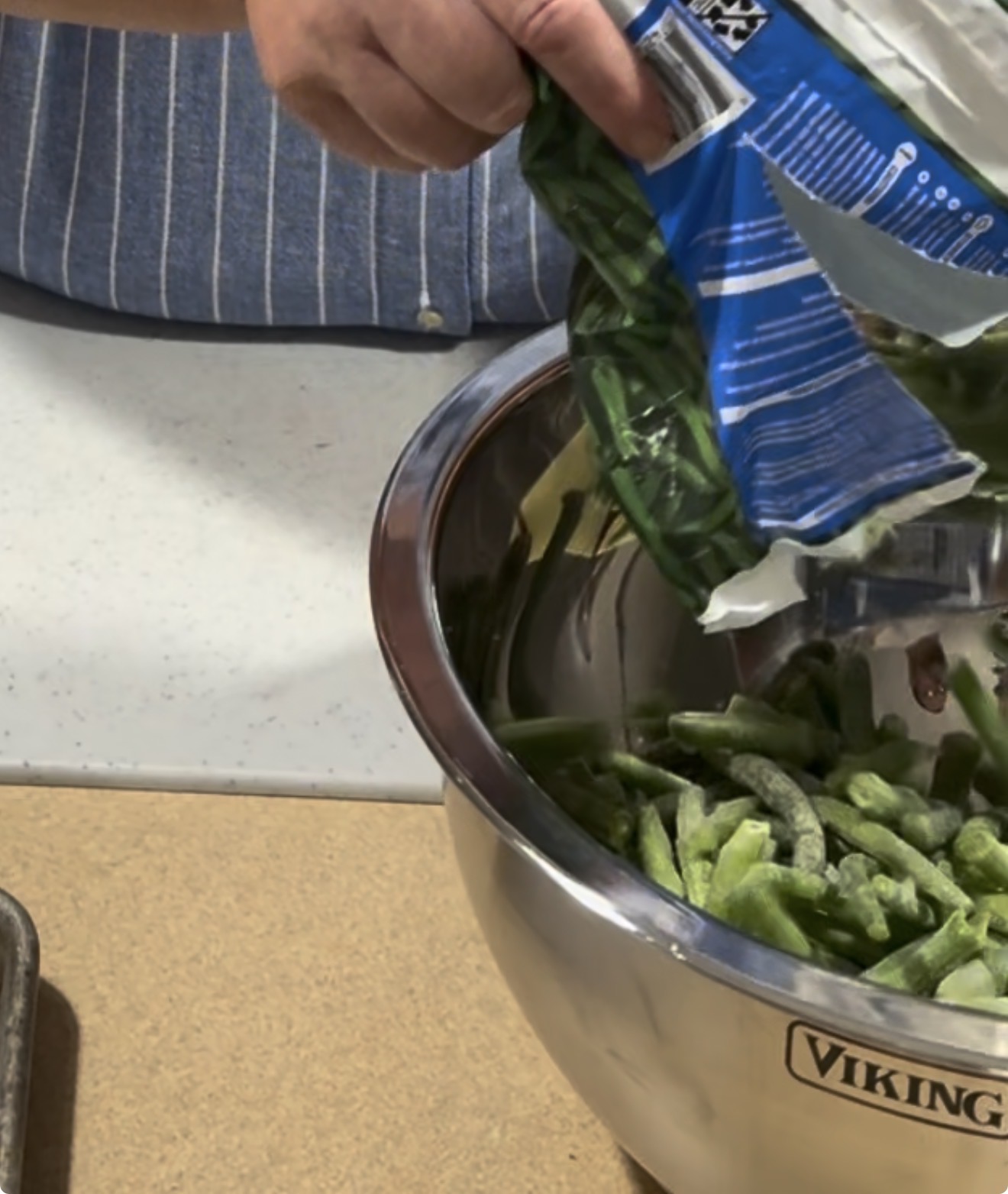 A person pours frozen green beans from a blue bag into a large metal mixing bowl on a kitchen counter.