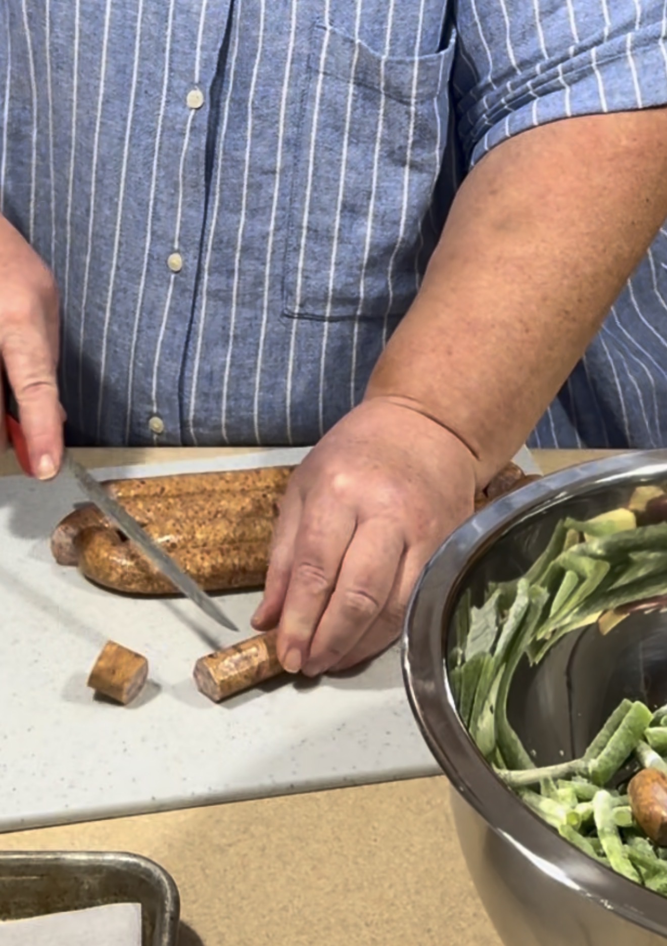 A person in a blue striped shirt slices sausage on a white cutting board next to a metal bowl filled with chopped vegetables.