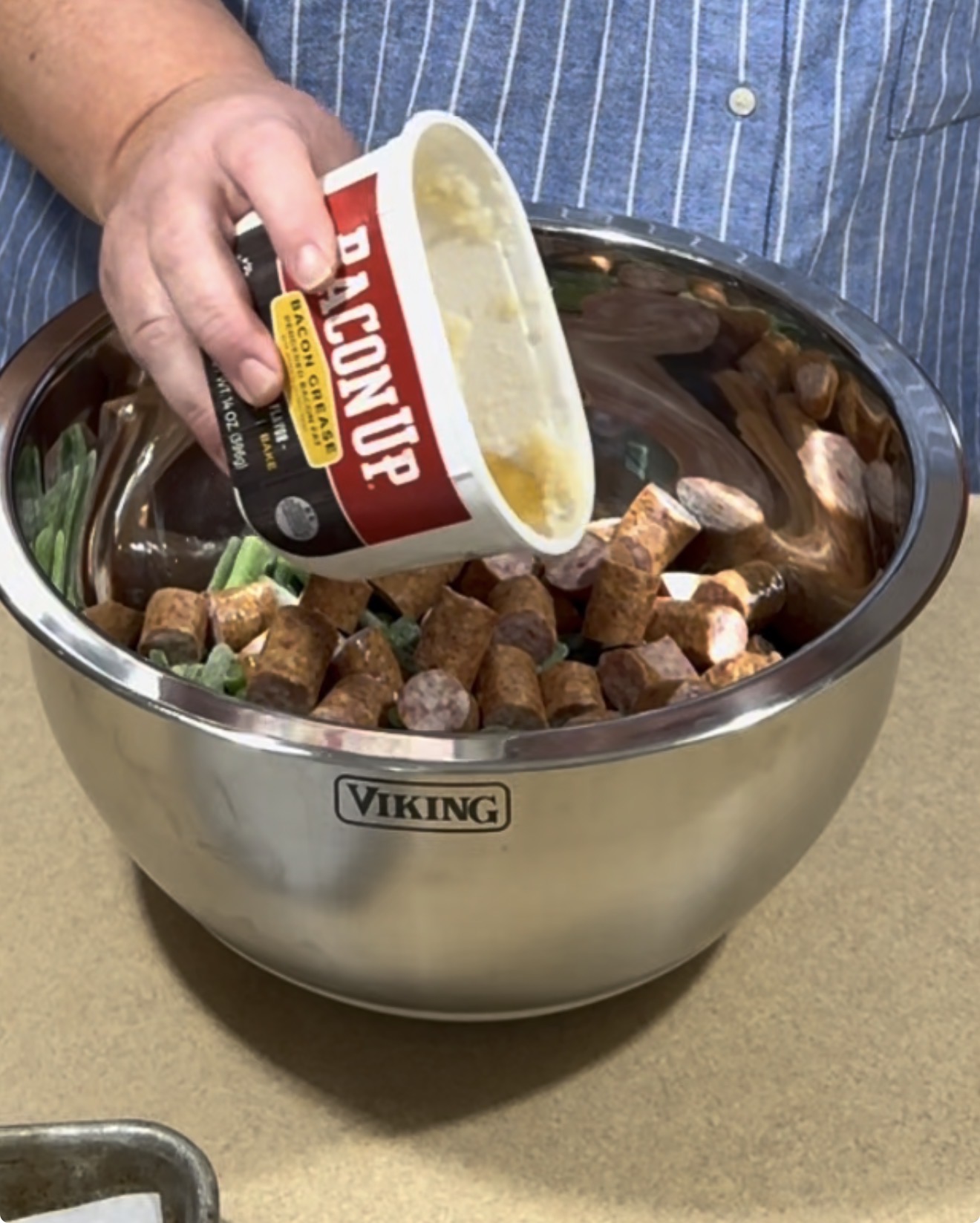 A person pours a container labeled Bacon Up into a large metal bowl filled with sliced sausage and green beans on a kitchen counter.