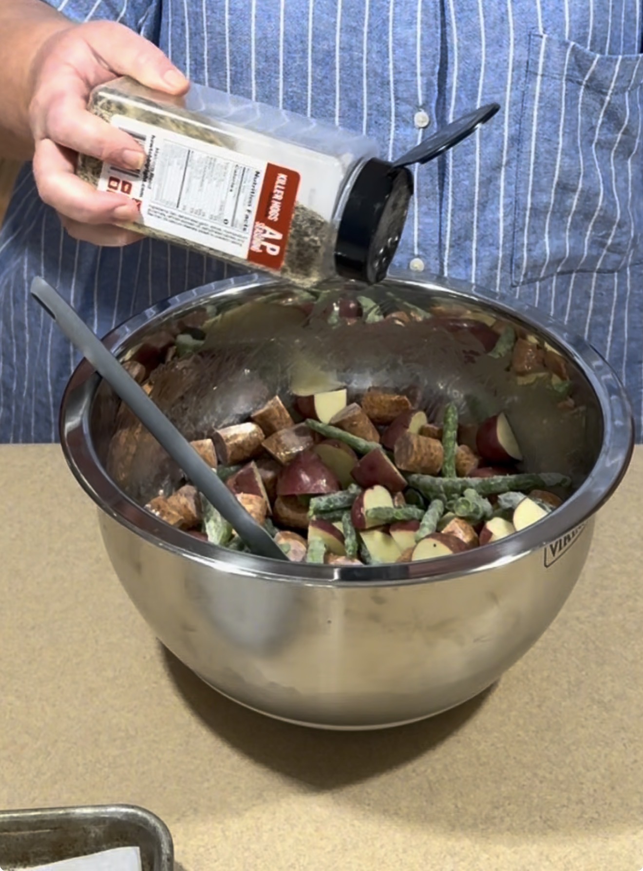 A person wearing a blue striped shirt sprinkles seasoning from a container into a large metal bowl filled with chopped potatoes, green beans, and sausage on a kitchen counter.