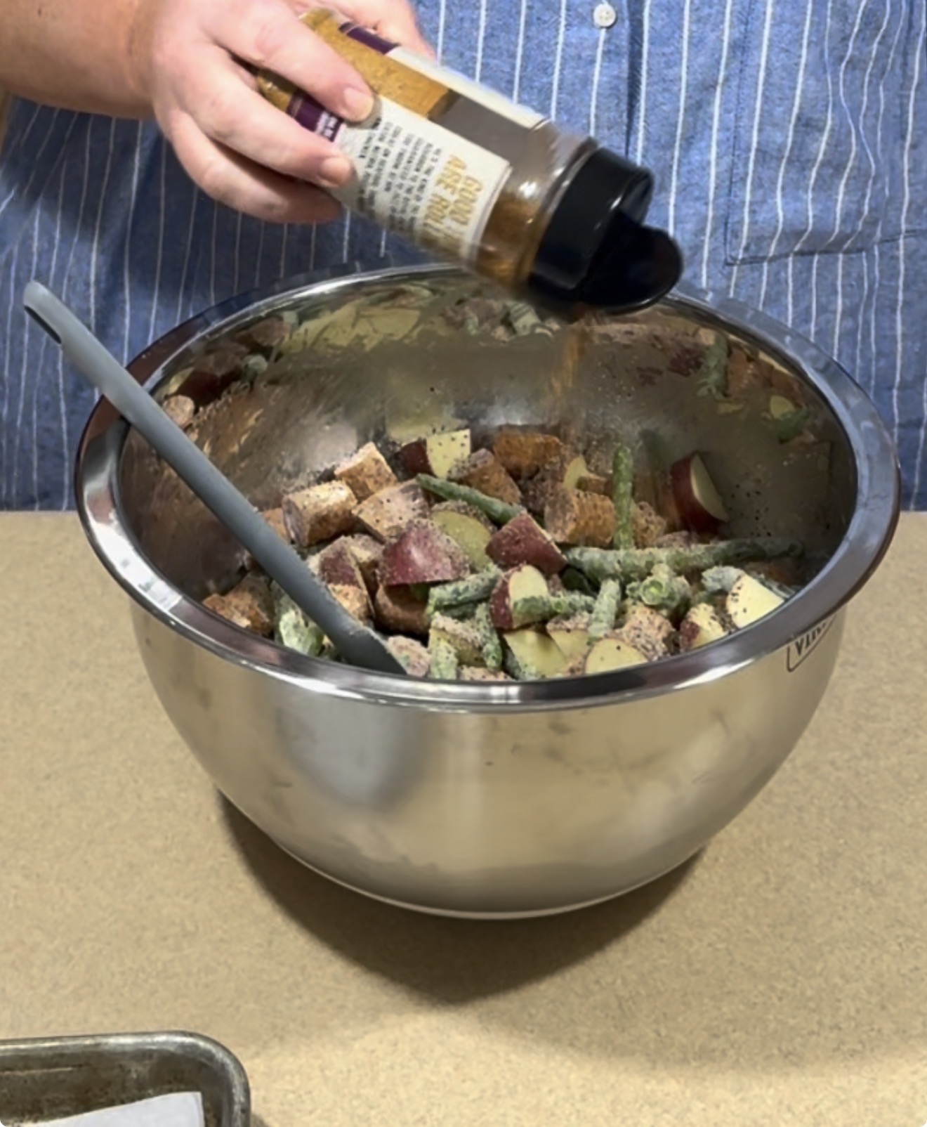 A person in a blue striped shirt sprinkles seasoning from a bottle into a large metal bowl filled with chopped potatoes and green beans, with a mixing spatula resting inside the bowl.