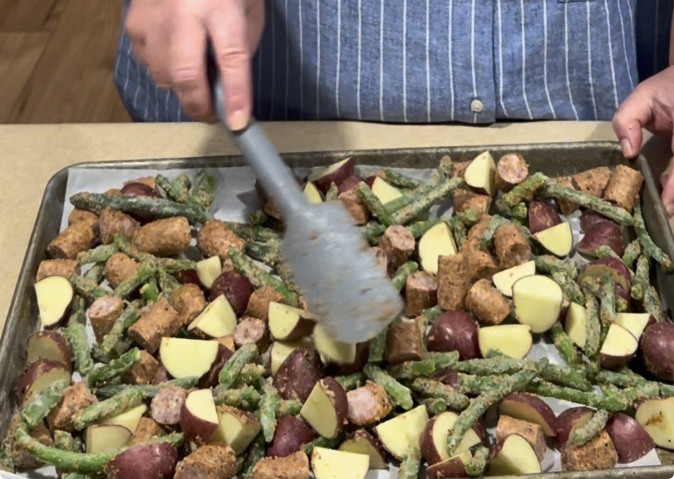 A person in a blue striped shirt uses tongs to mix green beans, sausage slices, and potato chunks on a baking sheet lined with parchment paper.