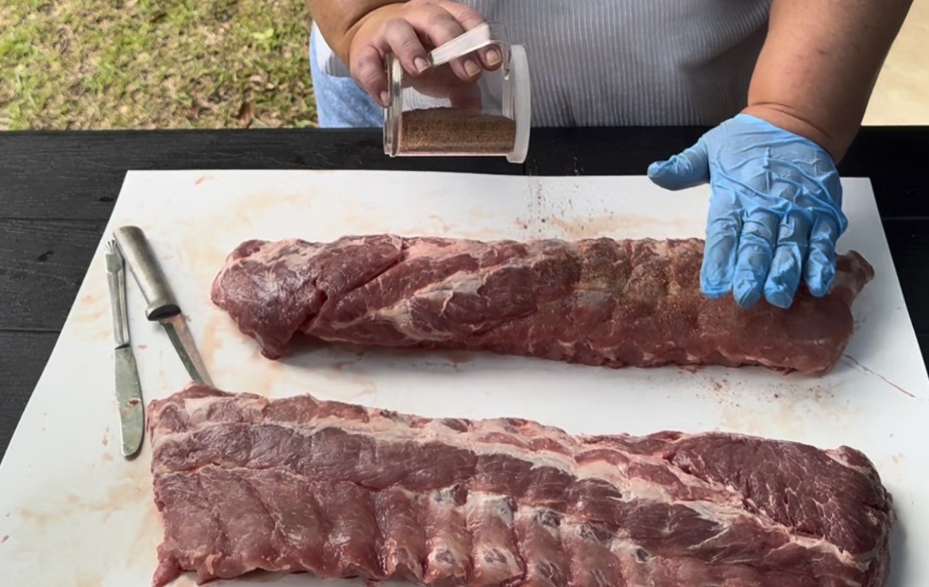 Person wearing a blue glove sprinkles seasoning onto raw racks of ribs on a white cutting board outside. Two knives are placed nearby on the black table.