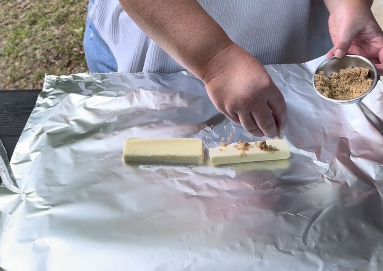 A person sprinkles brown sugar onto two sticks of butter placed on a sheet of aluminum foil outdoors.